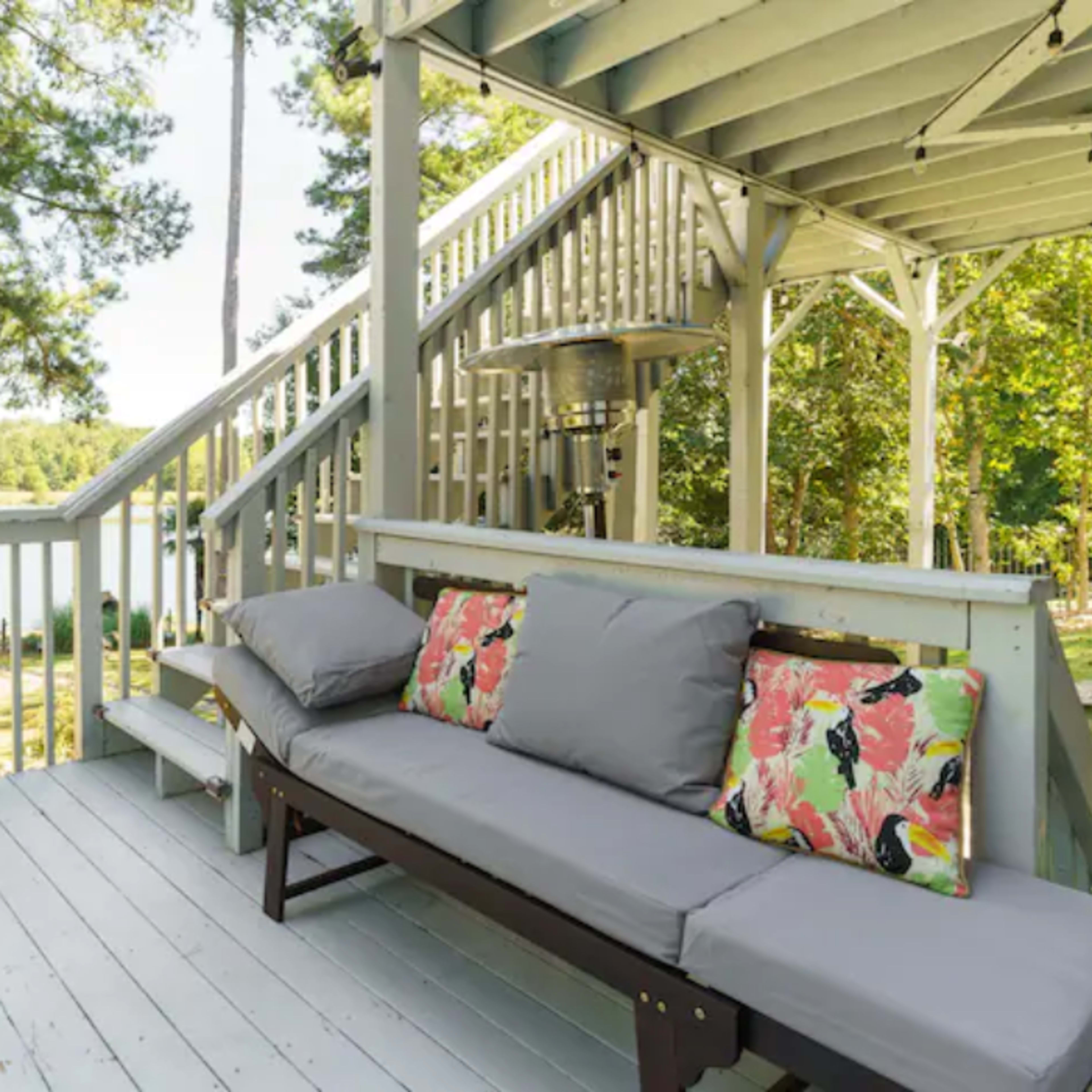 A wooden deck overlooking a lake, furnished with a couch adorned with colorful pillows and surrounded by greenery.