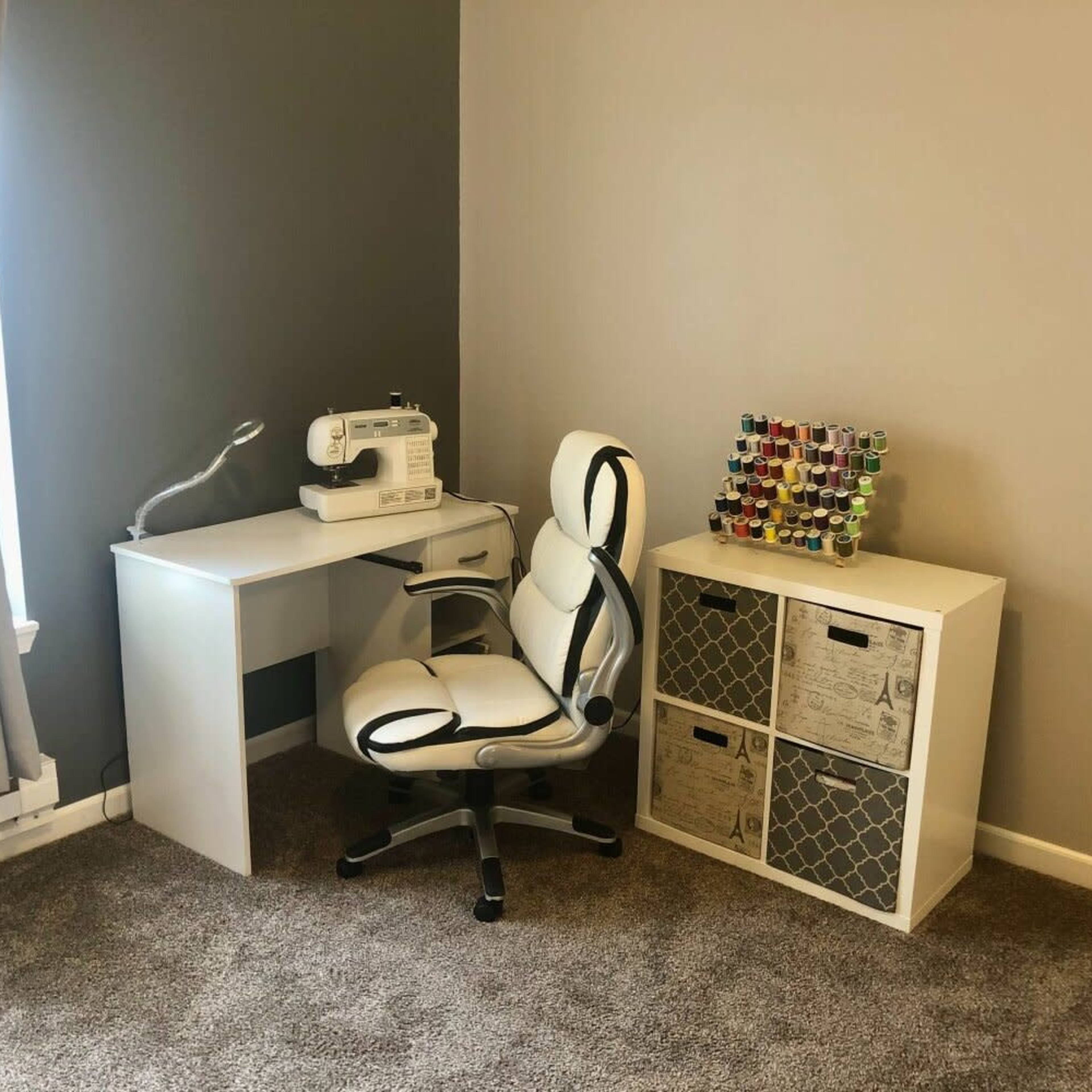A white sewing machine is placed on a minimalist desk next to a chair and a shelving unit filled with colorful spools of thread.