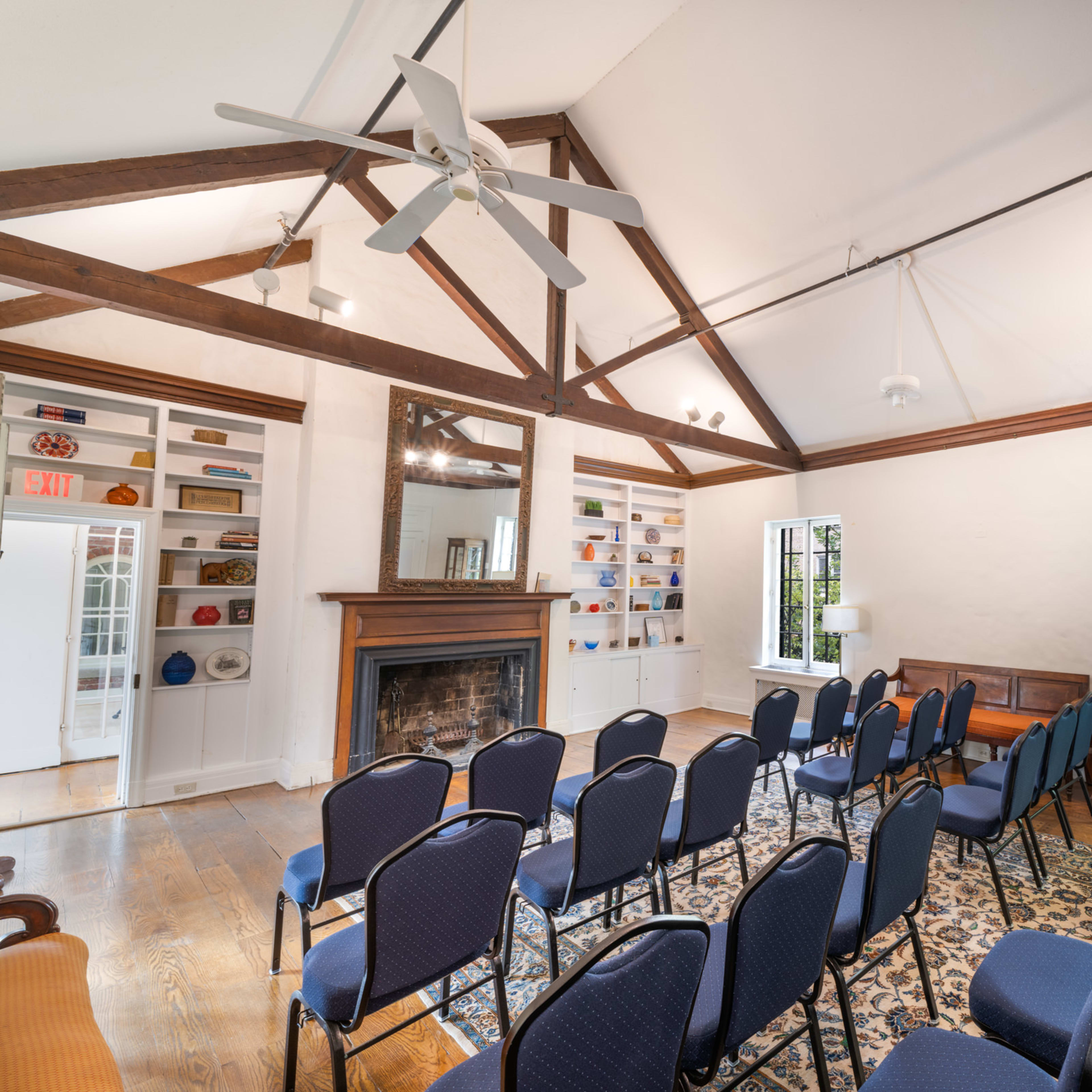A spacious room with high, beamed ceilings, featuring rows of blue chairs facing a fireplace and large windows.