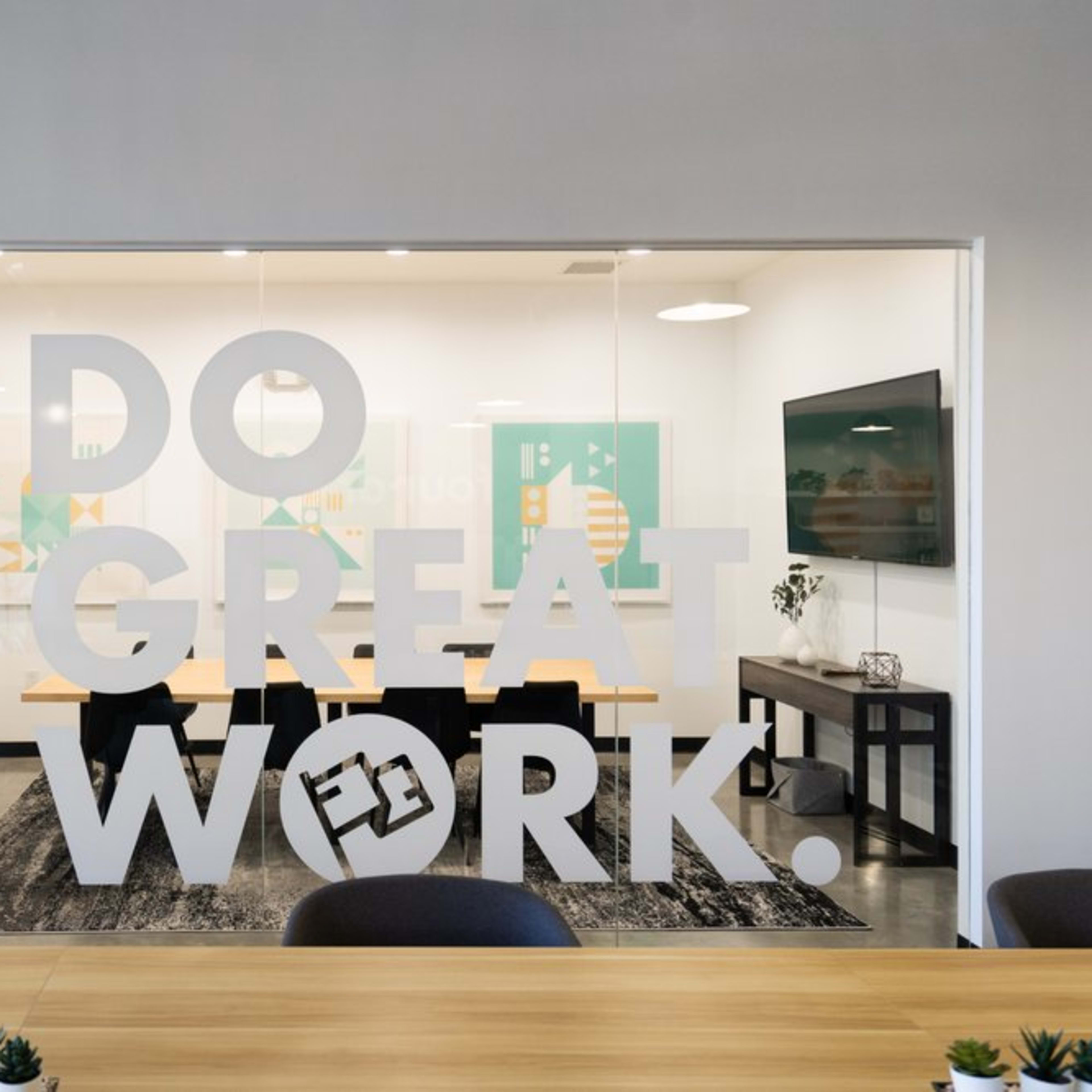 A glass-walled conference room features a modern table and chairs, with the phrase "DO GREAT WORK!" prominently displayed on the window.