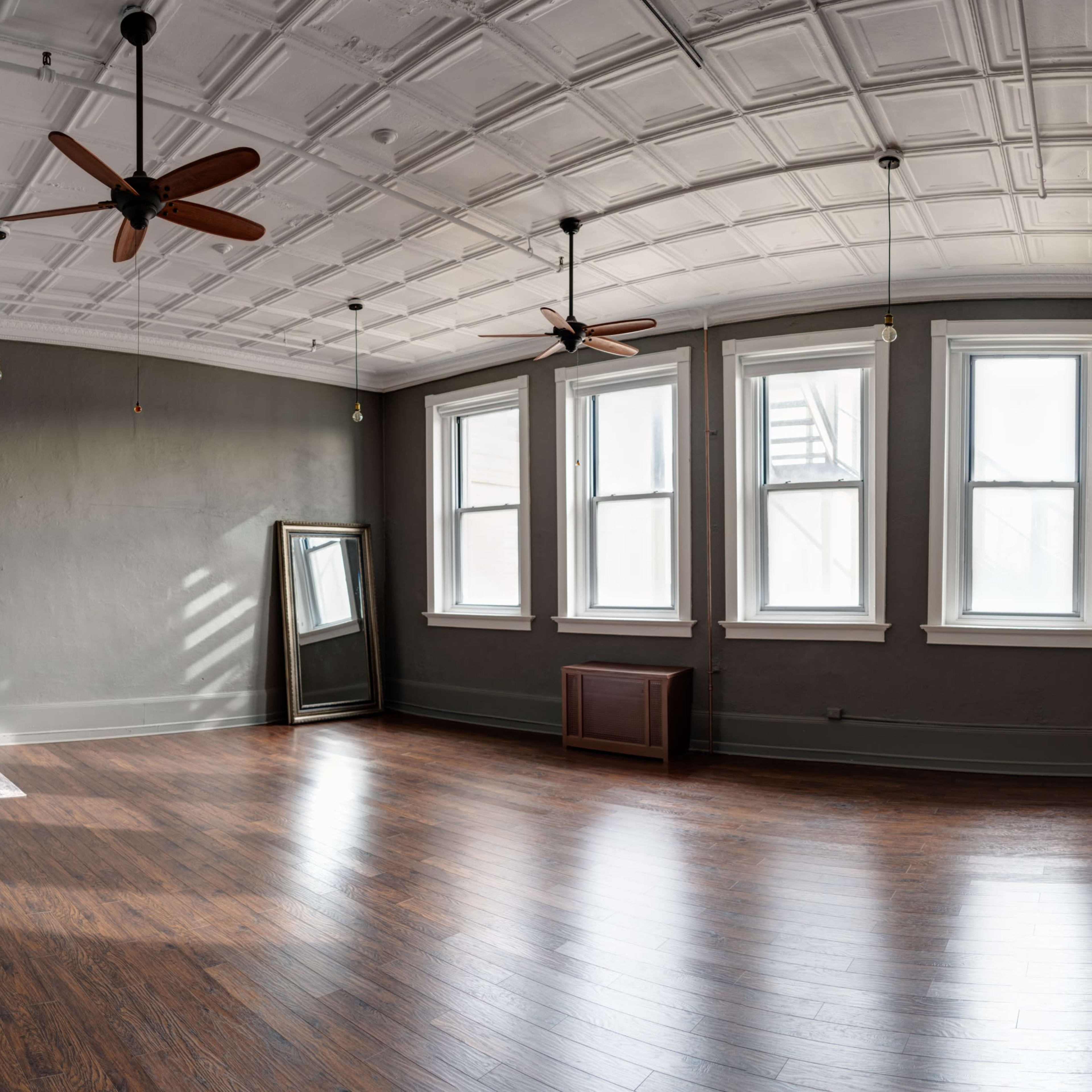 A spacious room with a wooden floor, large windows, a mirror, and ceiling fans, featuring a minimalistic design.