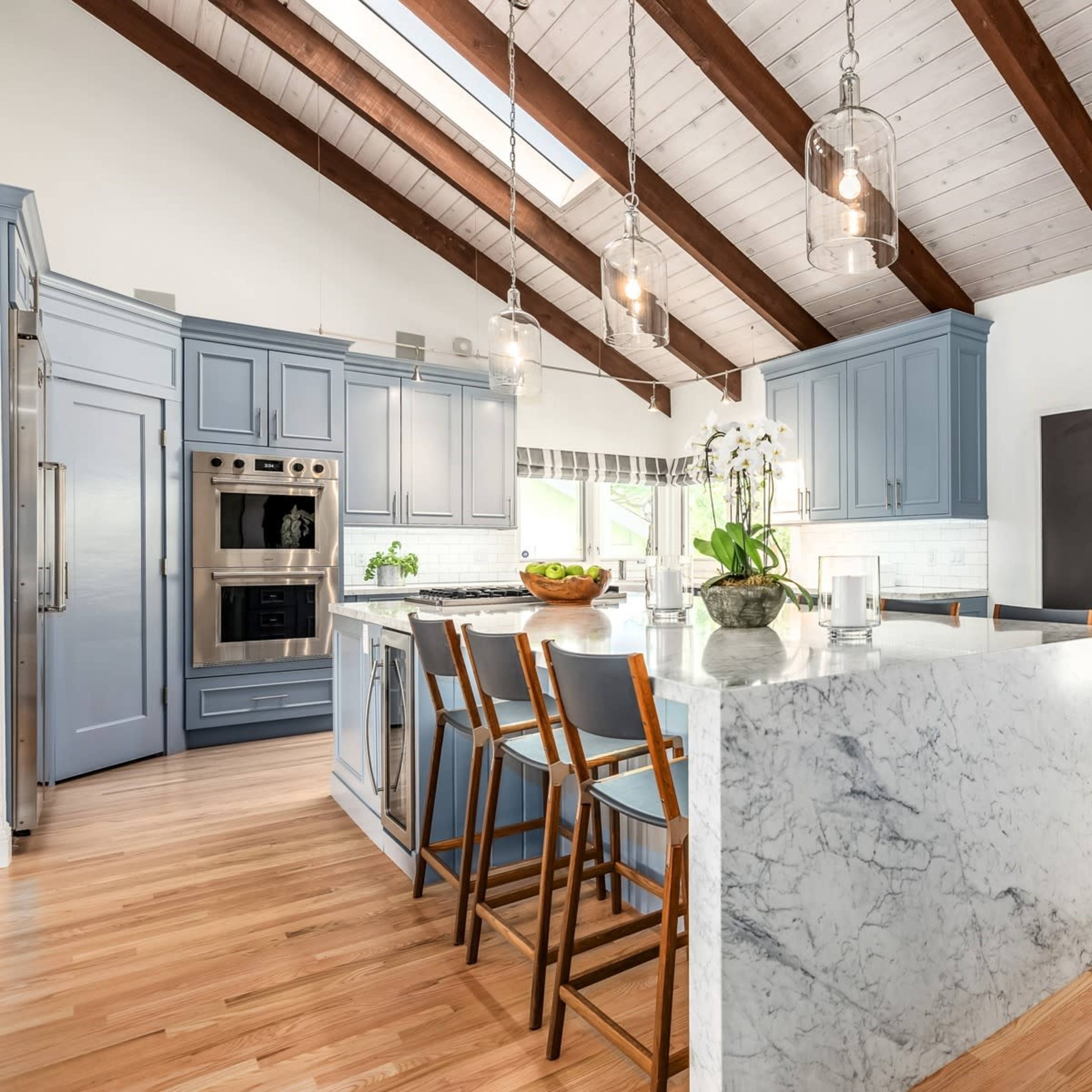 The kitchen features blue cabinetry, a marble island, and wooden beams, with large windows providing natural light.