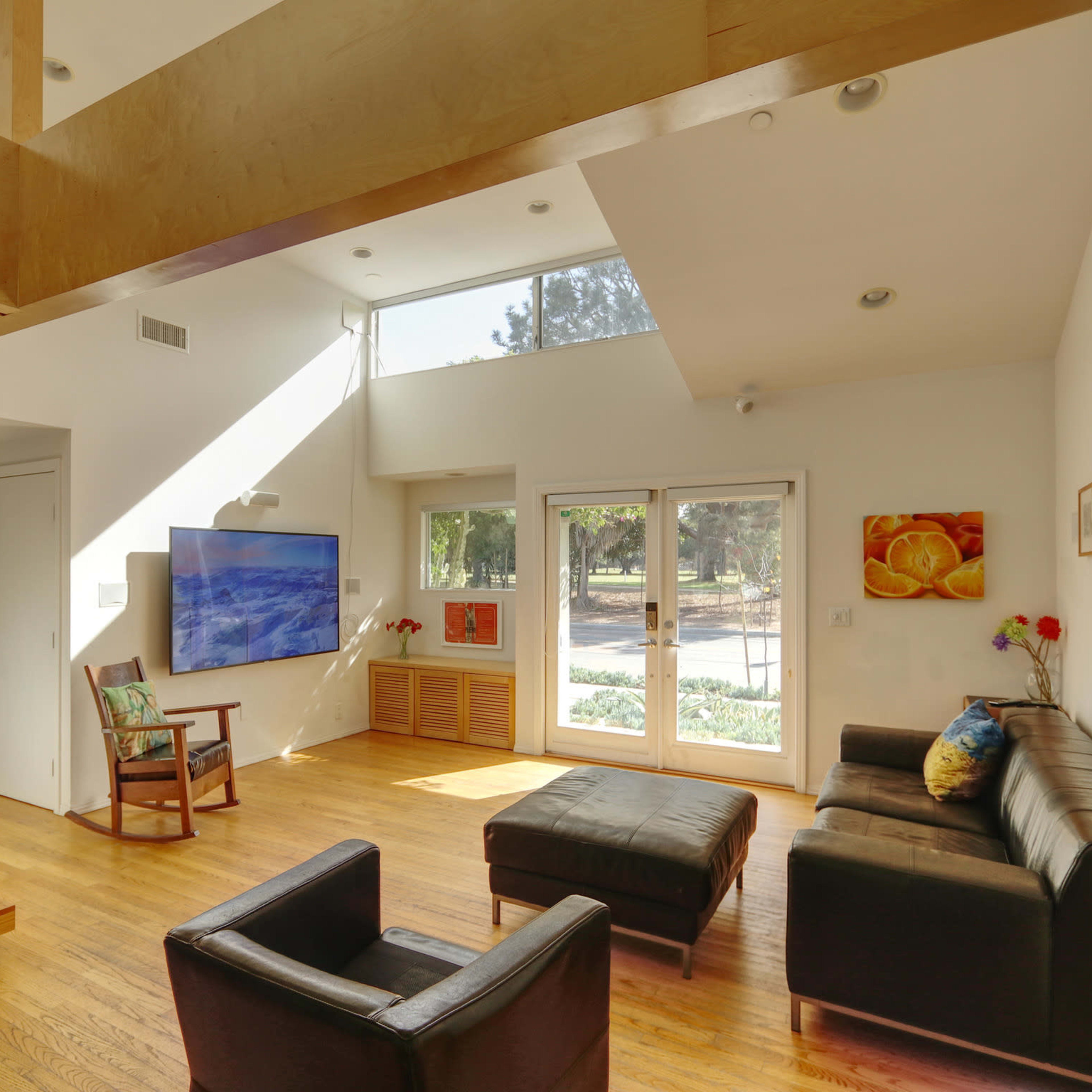 A bright living room with a minimalist design featuring a sectional sofa, two chairs, a television on the wall, and large windows that provide natural light.