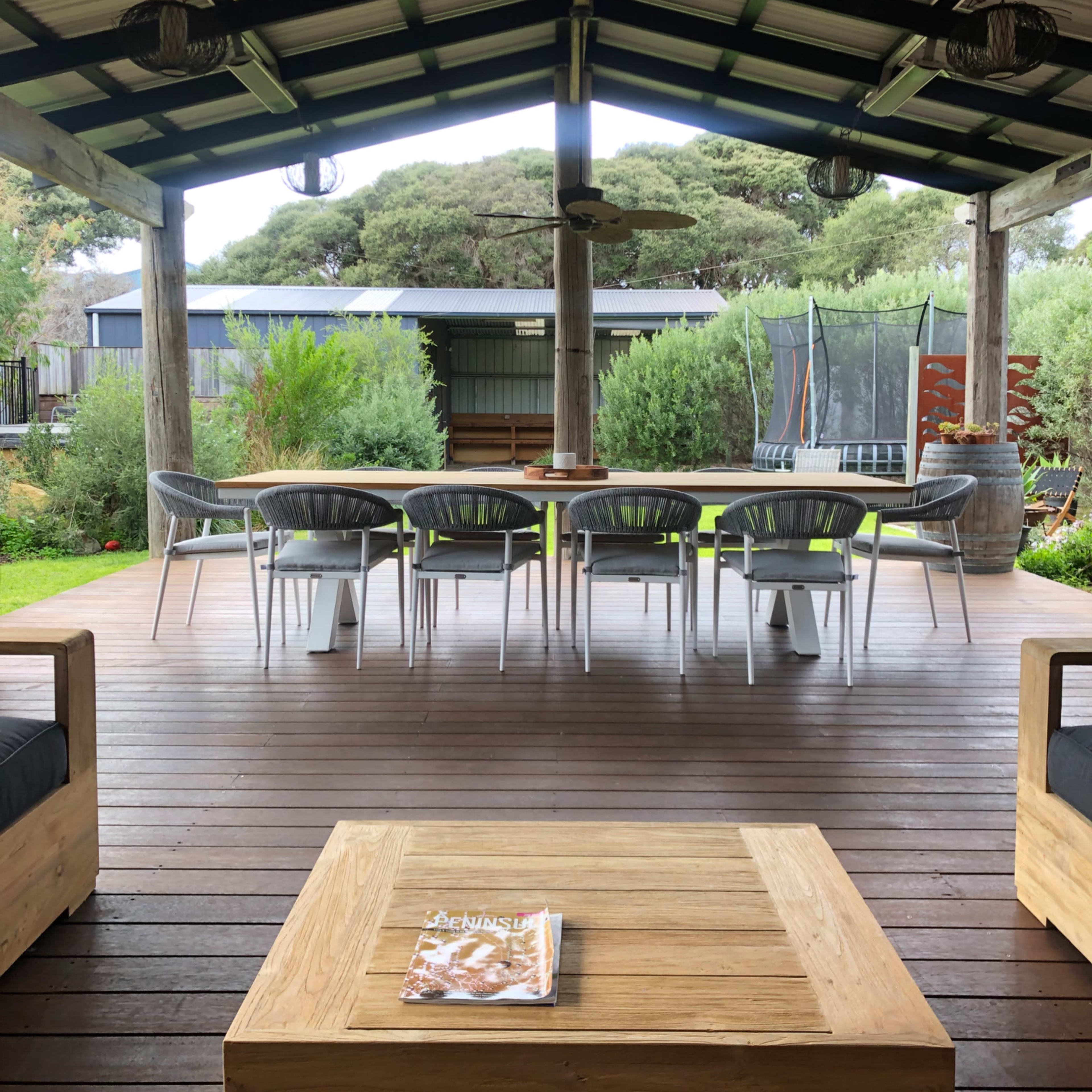 A spacious outdoor seating area features a large table surrounded by chairs under a covered wooden patio, with greenery and a trampoline visible in the background.