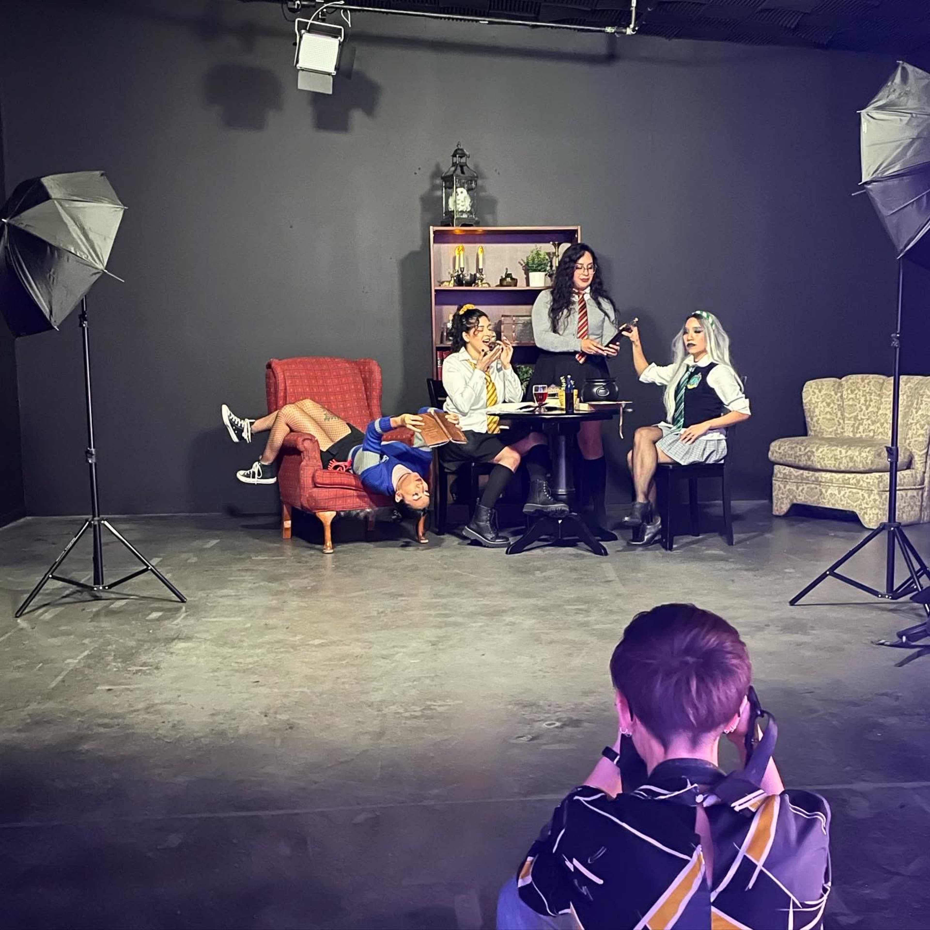 Four individuals are posing for a photo in a studio setting, with two sitting on couches and one standing, while another reclines on the floor.