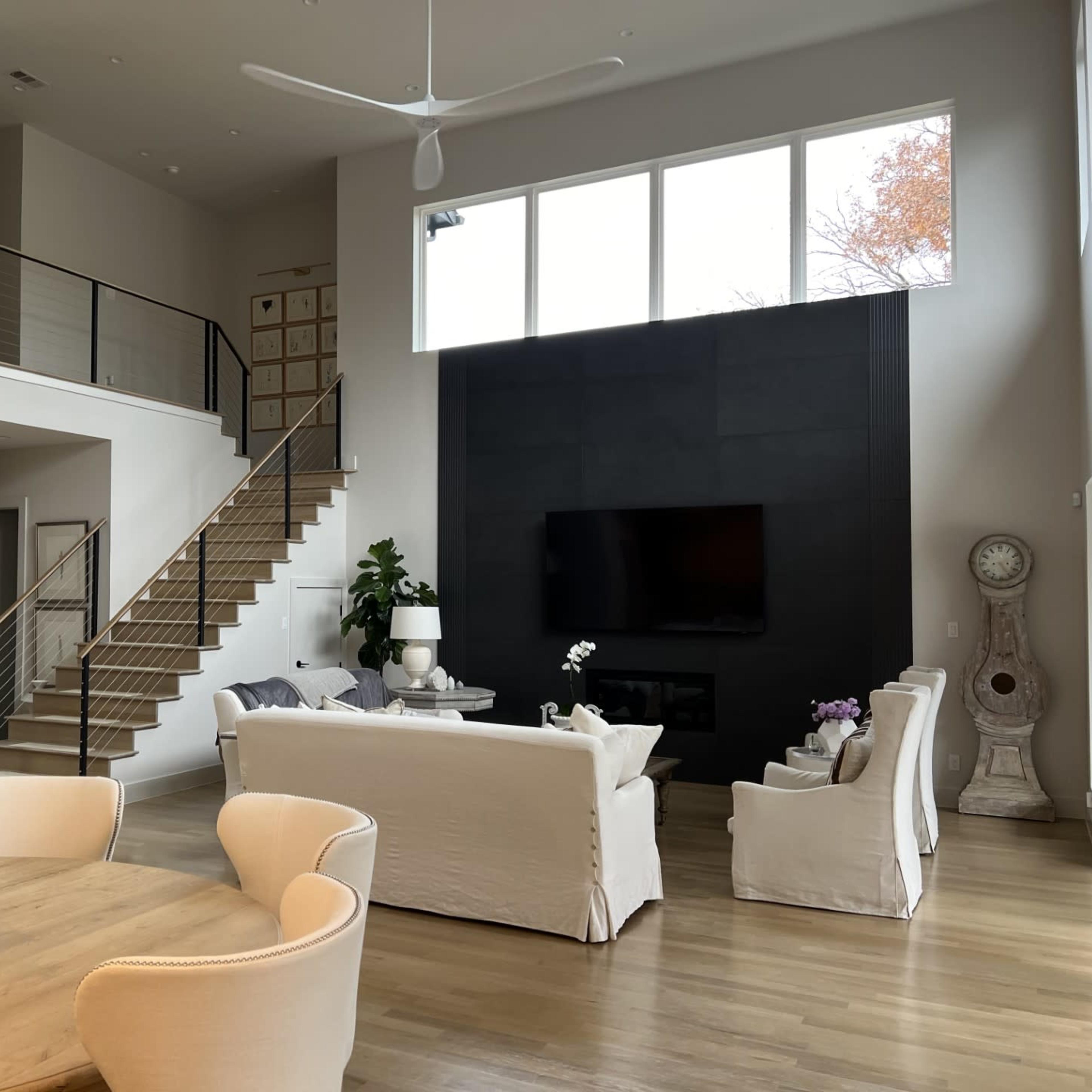 A modern open-concept living room with a staircase, a large wall-mounted TV, and light-colored furniture around a wooden dining table.