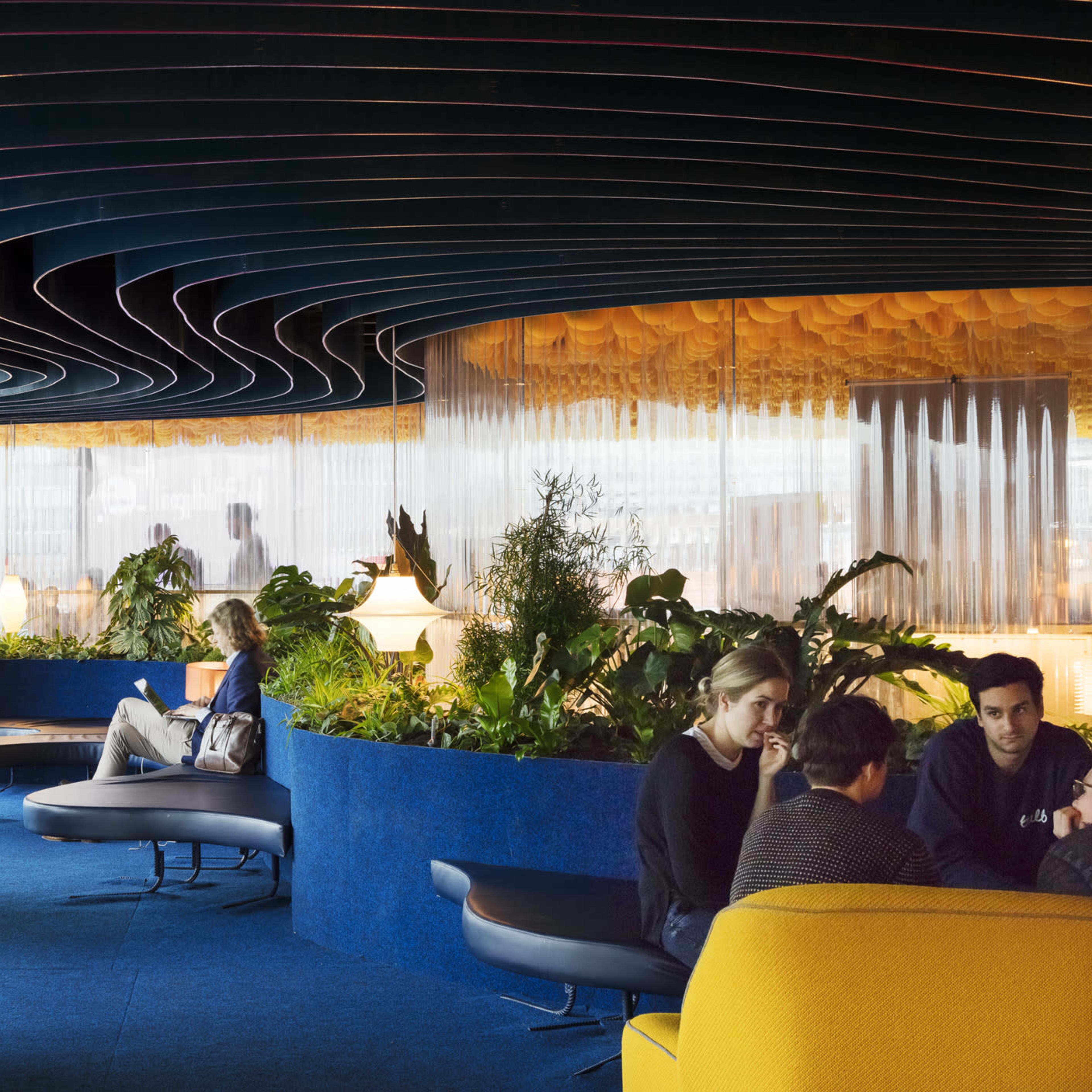 A modern interior space featuring curved ceiling design, various seating arrangements, and greenery, with people engaged in conversation and relaxation.