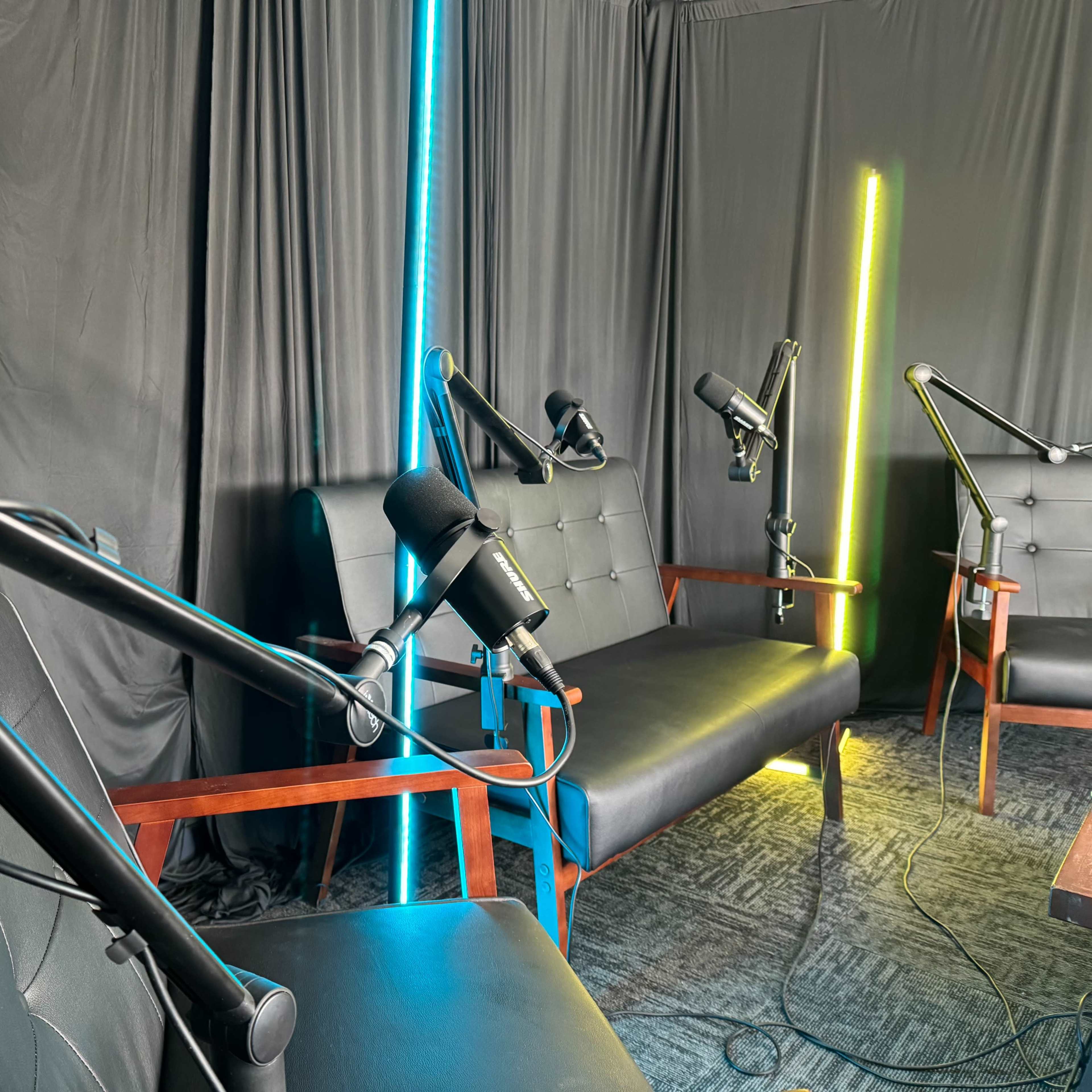 A recording studio with two leather sofas and microphone stands positioned in front of black drapes and illuminated by colored lights.