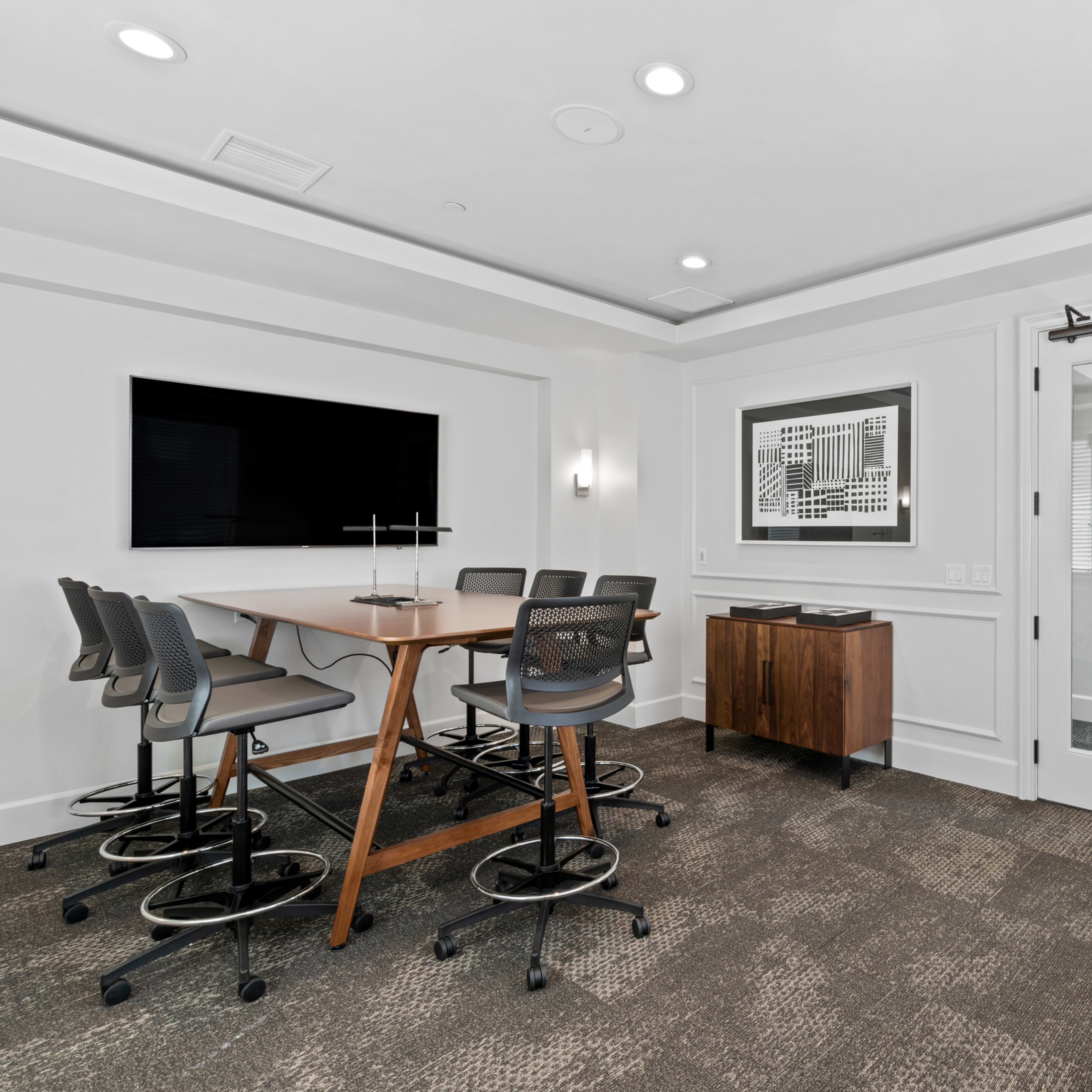 A modern conference room with a large table, several chairs, a wall-mounted TV, and glass doors leading to another area.