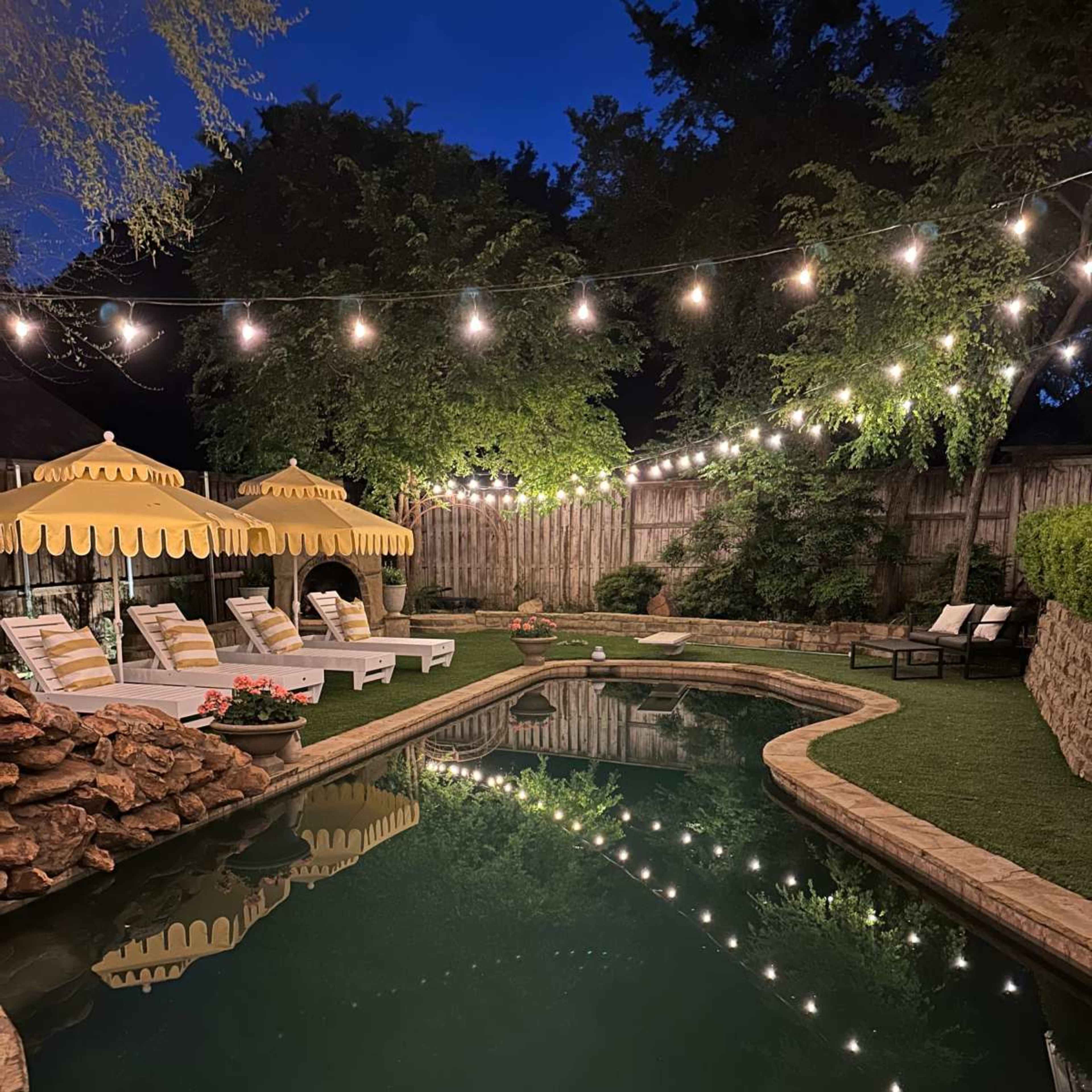 A backyard pool area features lounge chairs and umbrellas, illuminated by string lights along the water's edge.