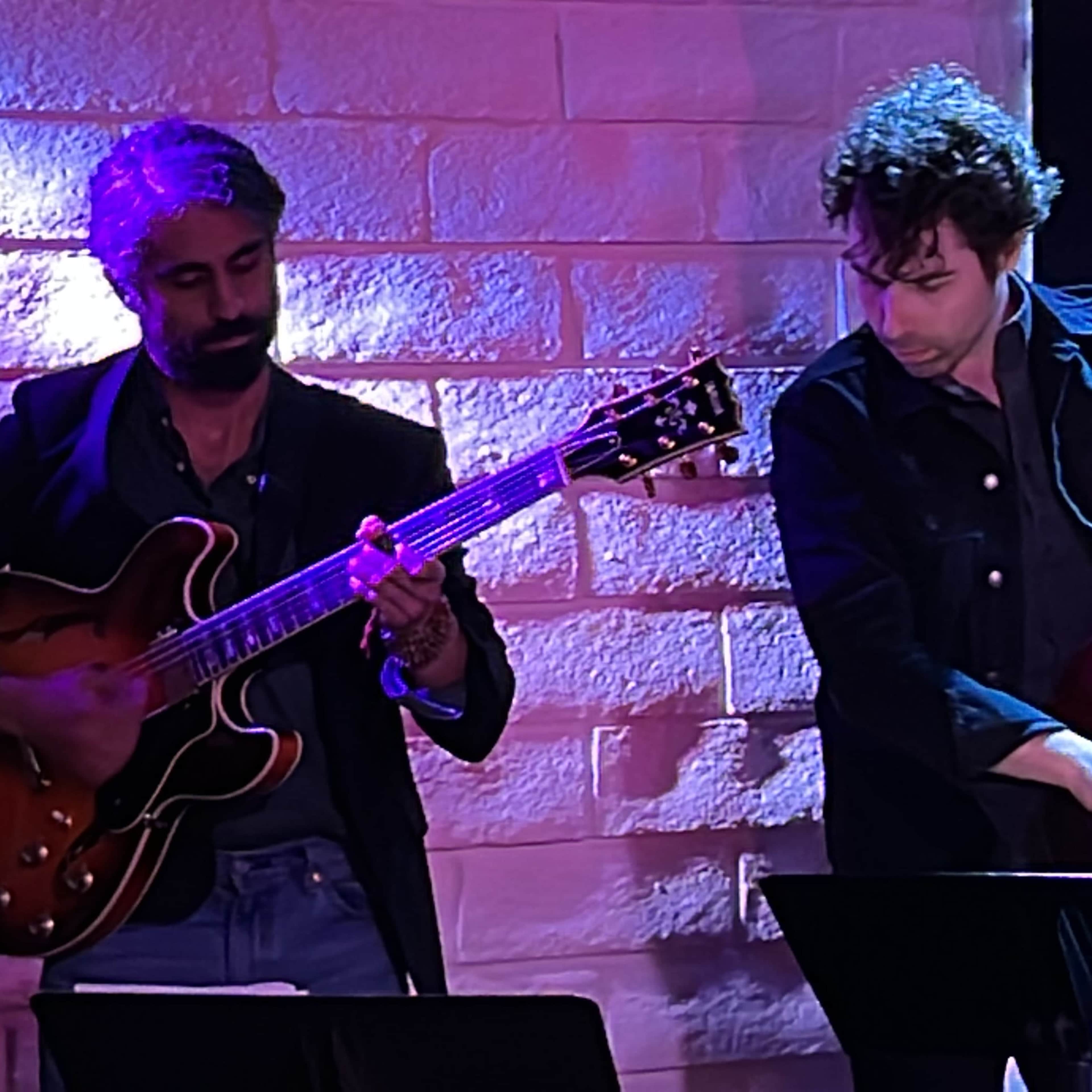 Two musicians perform live on stage, one playing an electric guitar and the other a double bass, against a backdrop of illuminated brick walls.