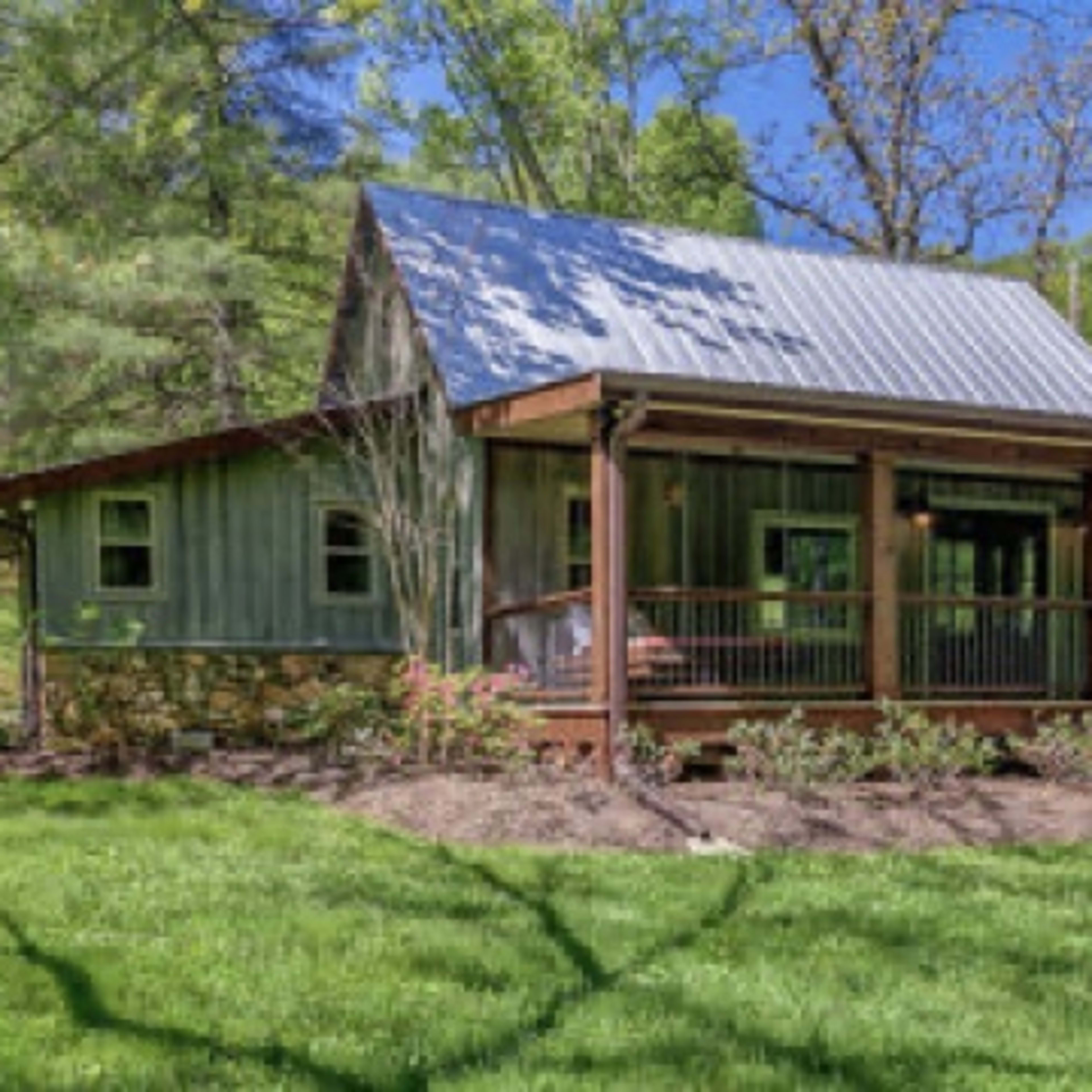 A rustic house with a metal roof and a wooden porch is surrounded by lush green grass and trees.