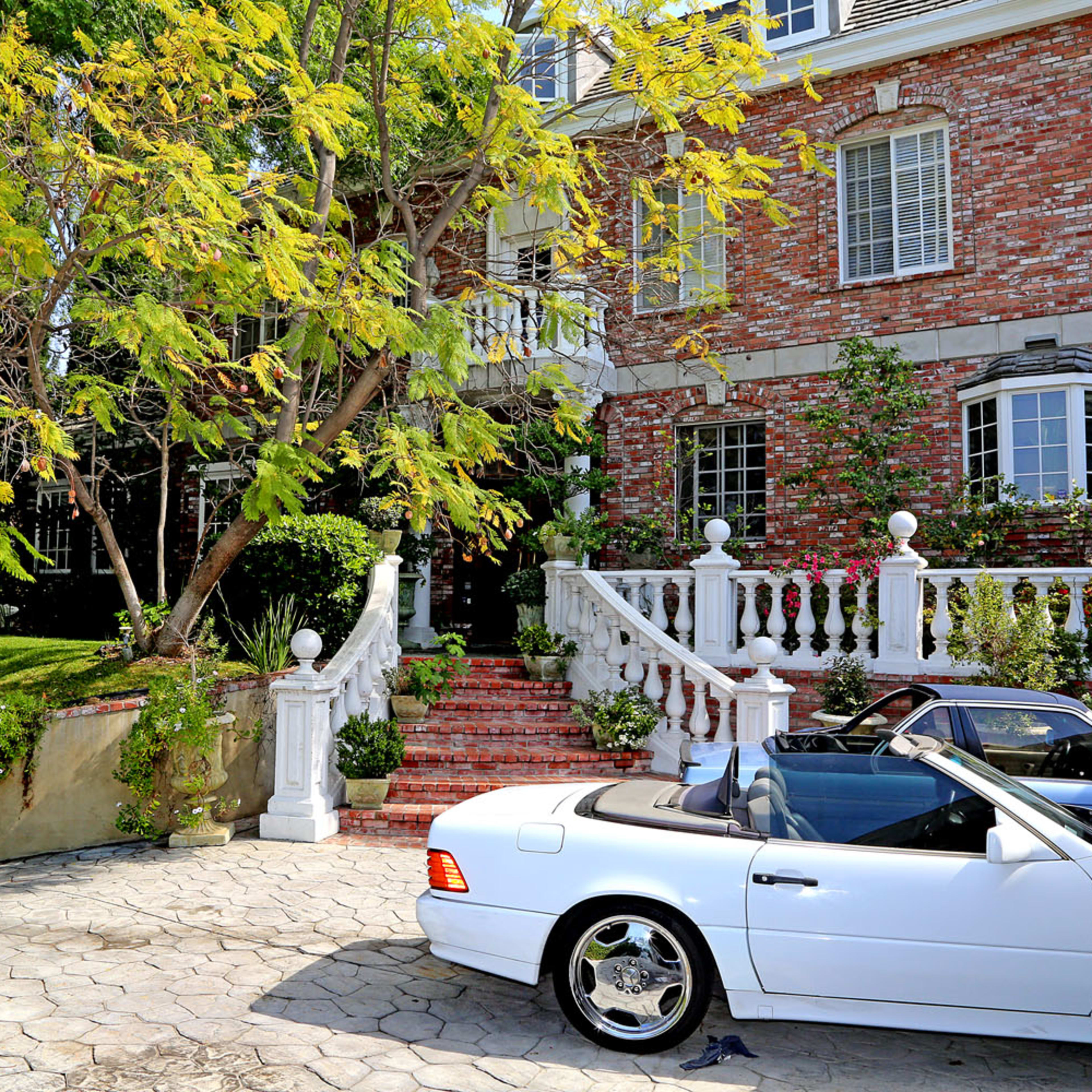 A white convertible car is parked in front of a brick mansion with a landscaped yard and a staircase flanked by white columns.