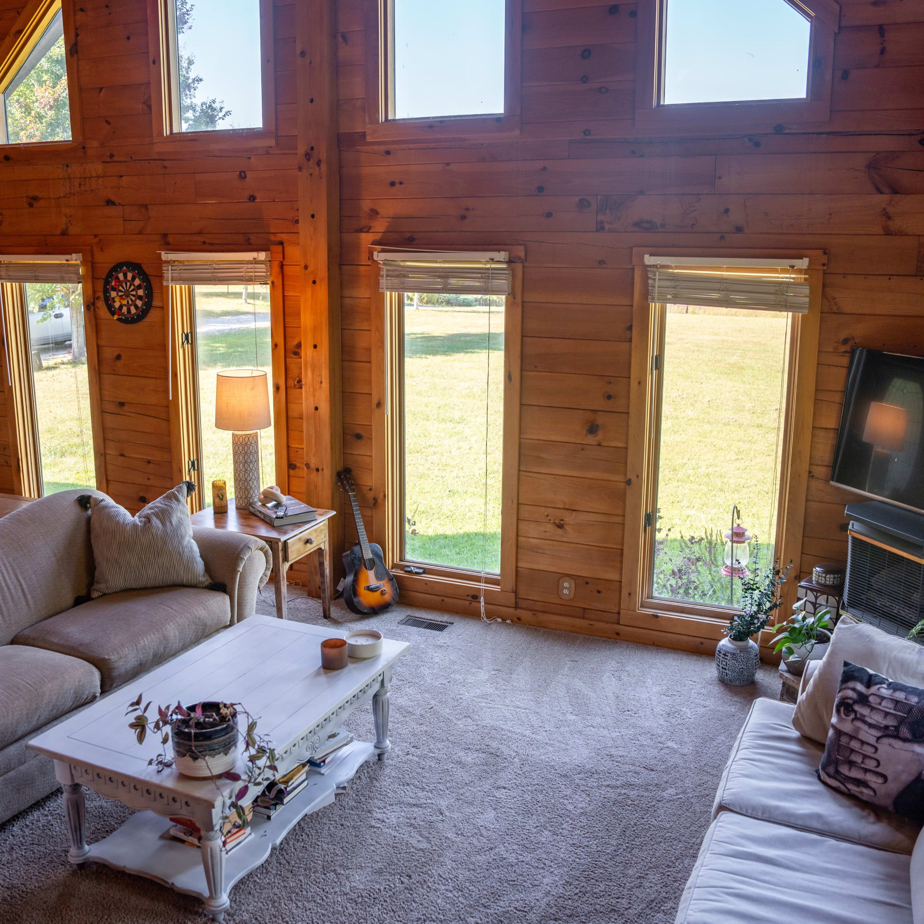 A wooden cabin living room with large windows, a coffee table, two sofas, and a guitar resting in the corner.