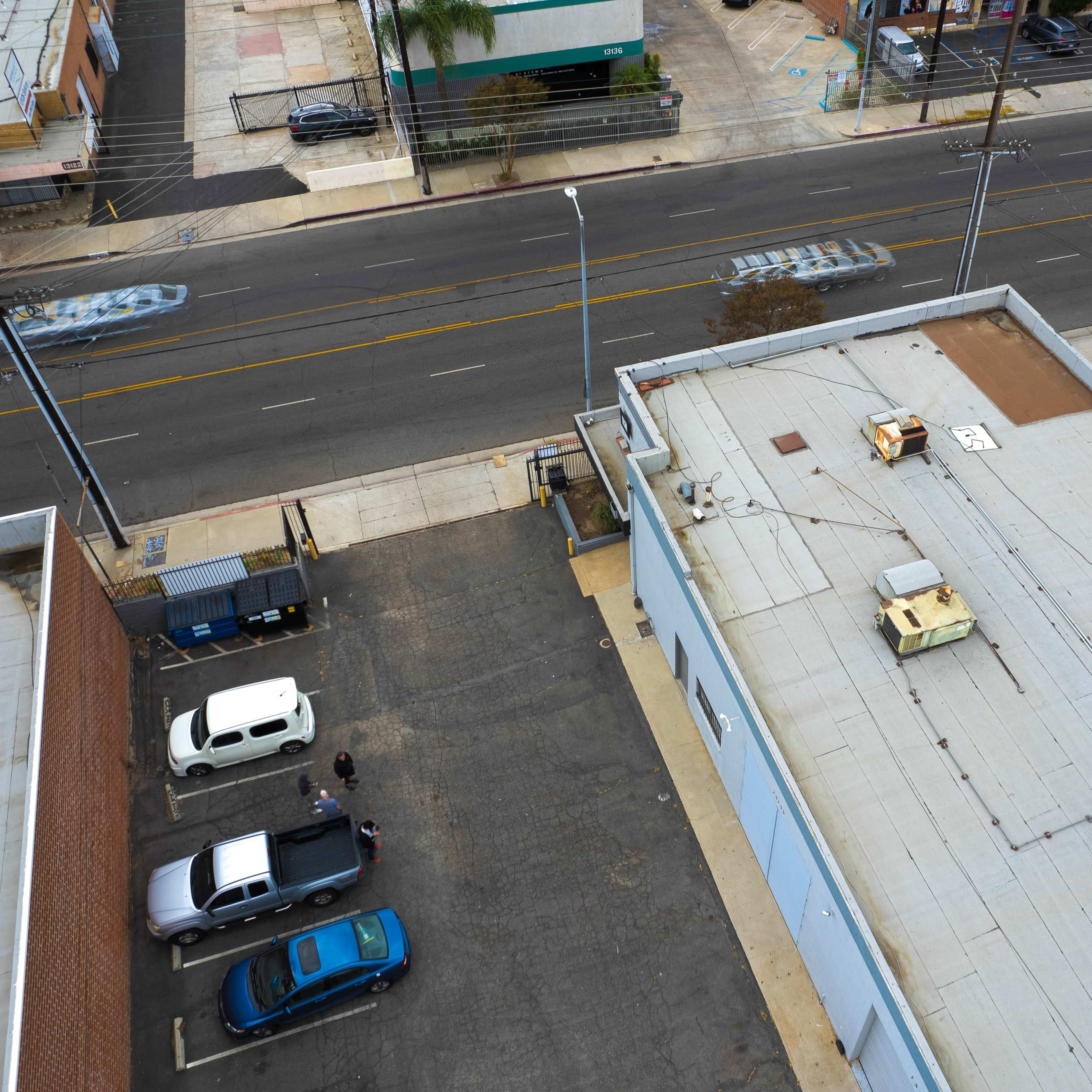 A bird's-eye view of a parking lot adjacent to a commercial building, with several parked cars and a street lined with buildings in the background.
