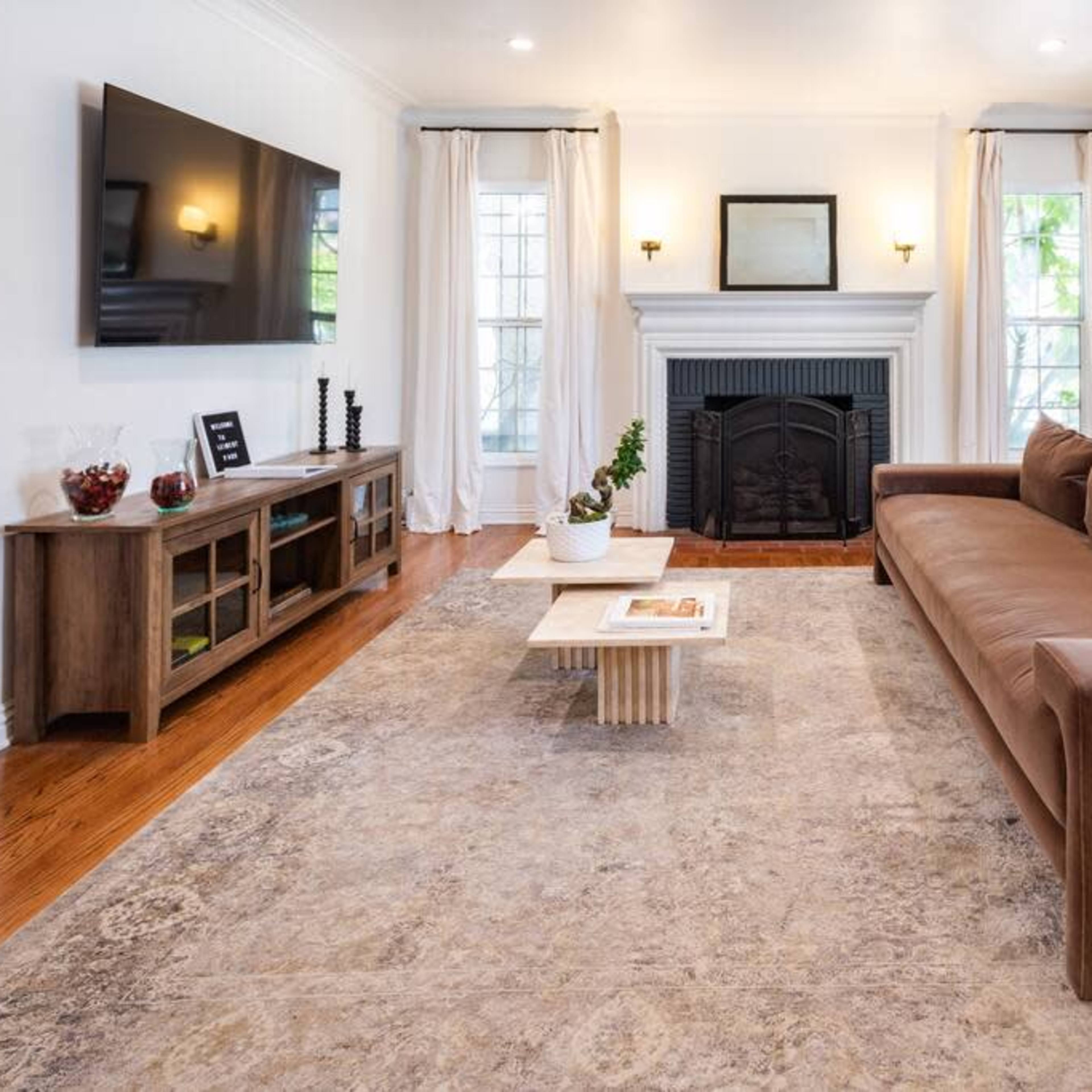 The living room features a brown sofa facing a mounted television and a fireplace, with a coffee table and a large area rug on polished wooden floors.