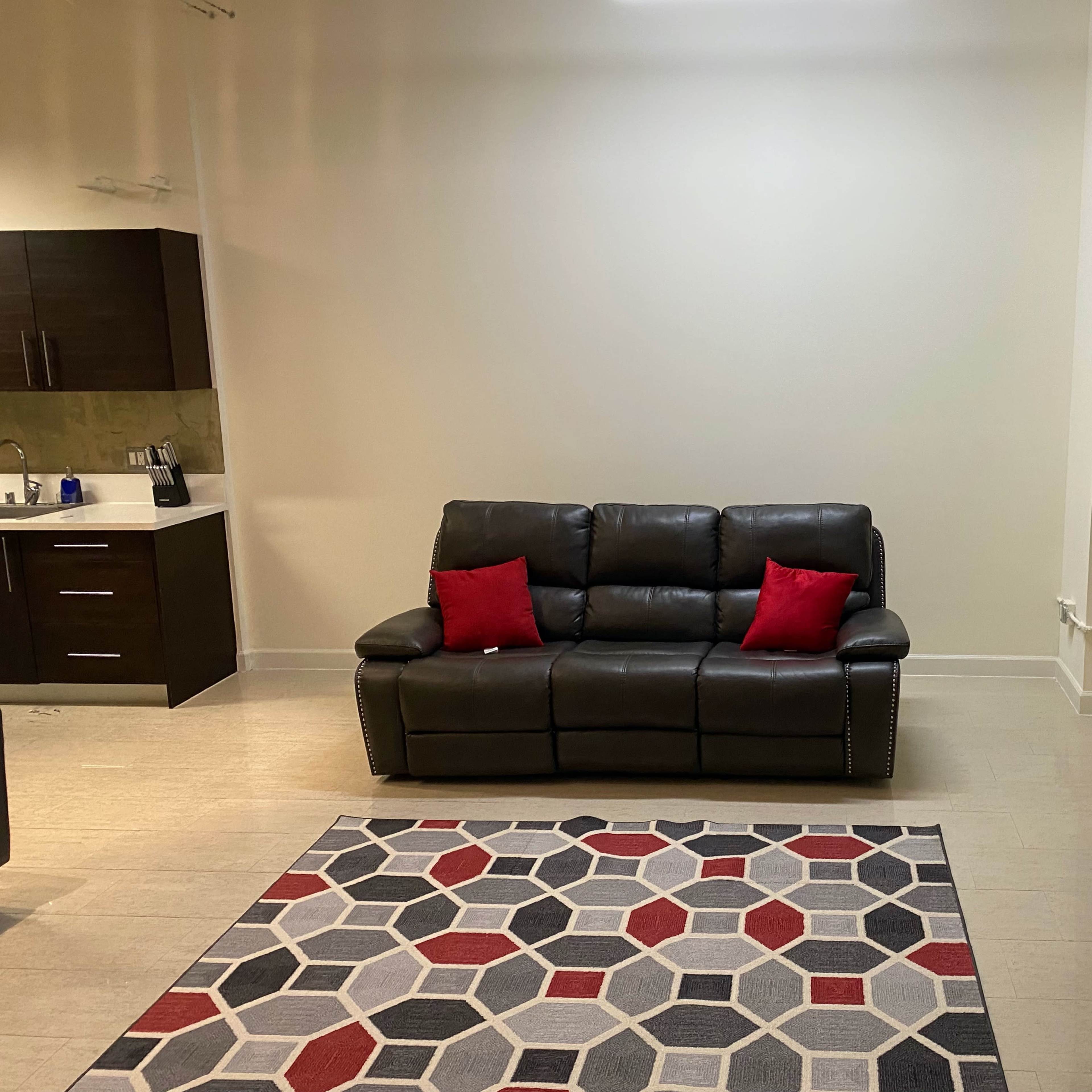A leather sofa with red decorative pillows sits in a spacious room featuring a geometric patterned rug and minimalistic decor.