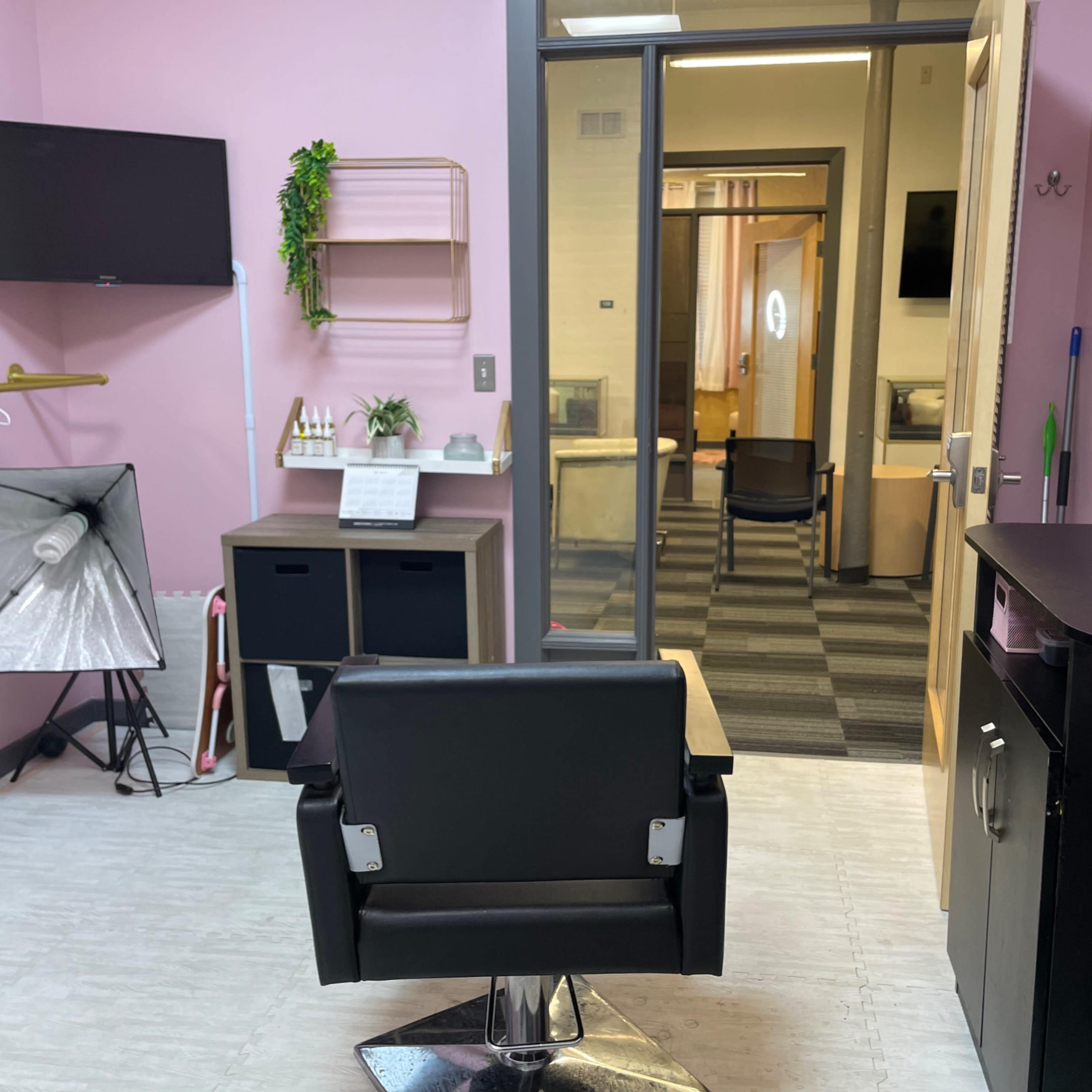 A brightly colored salon room with a black styling chair, a mirror, and storage units, leading into additional workspaces through an open door.