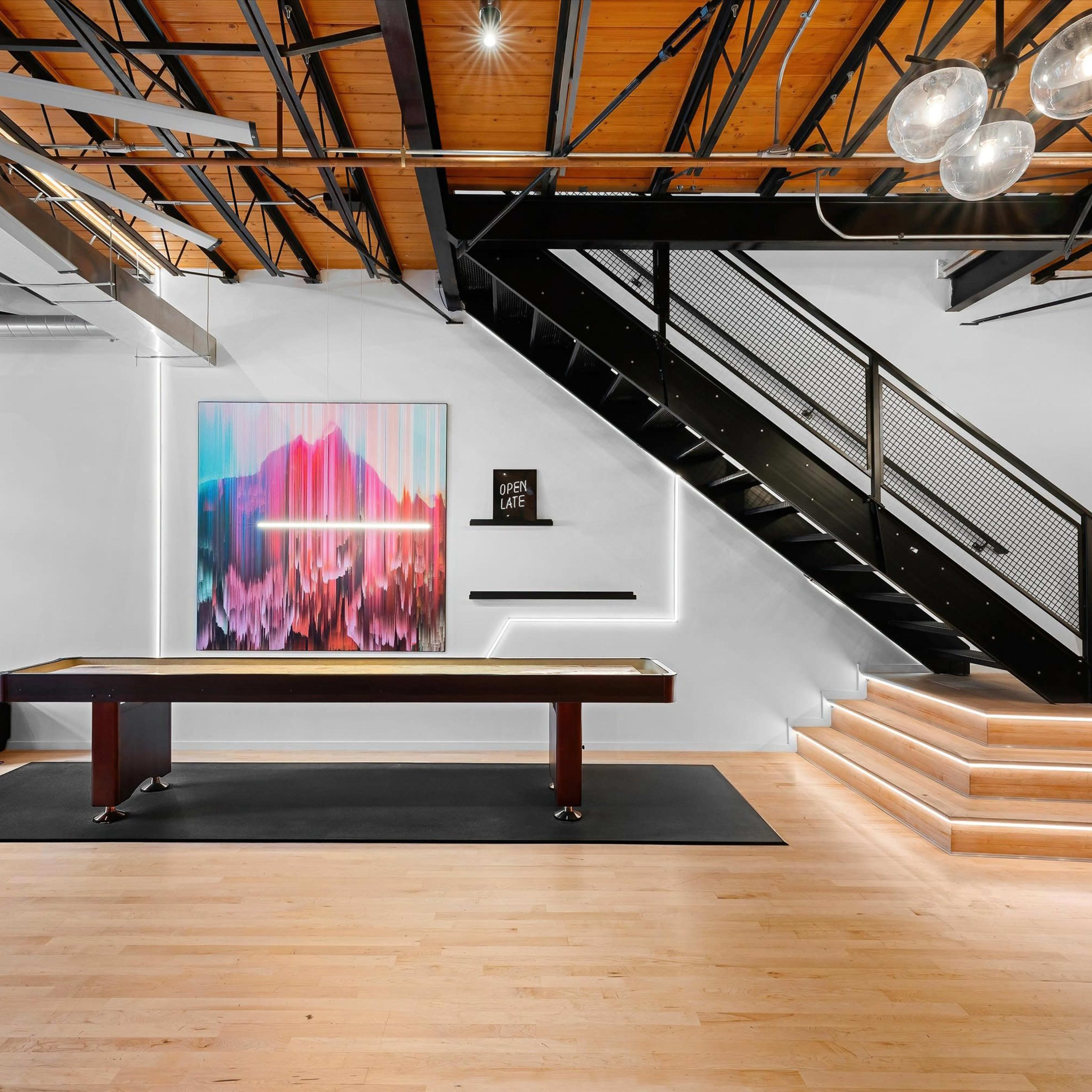 A modern interior space featuring a pool table, a staircase with black railings, and colorful artwork on the wall.