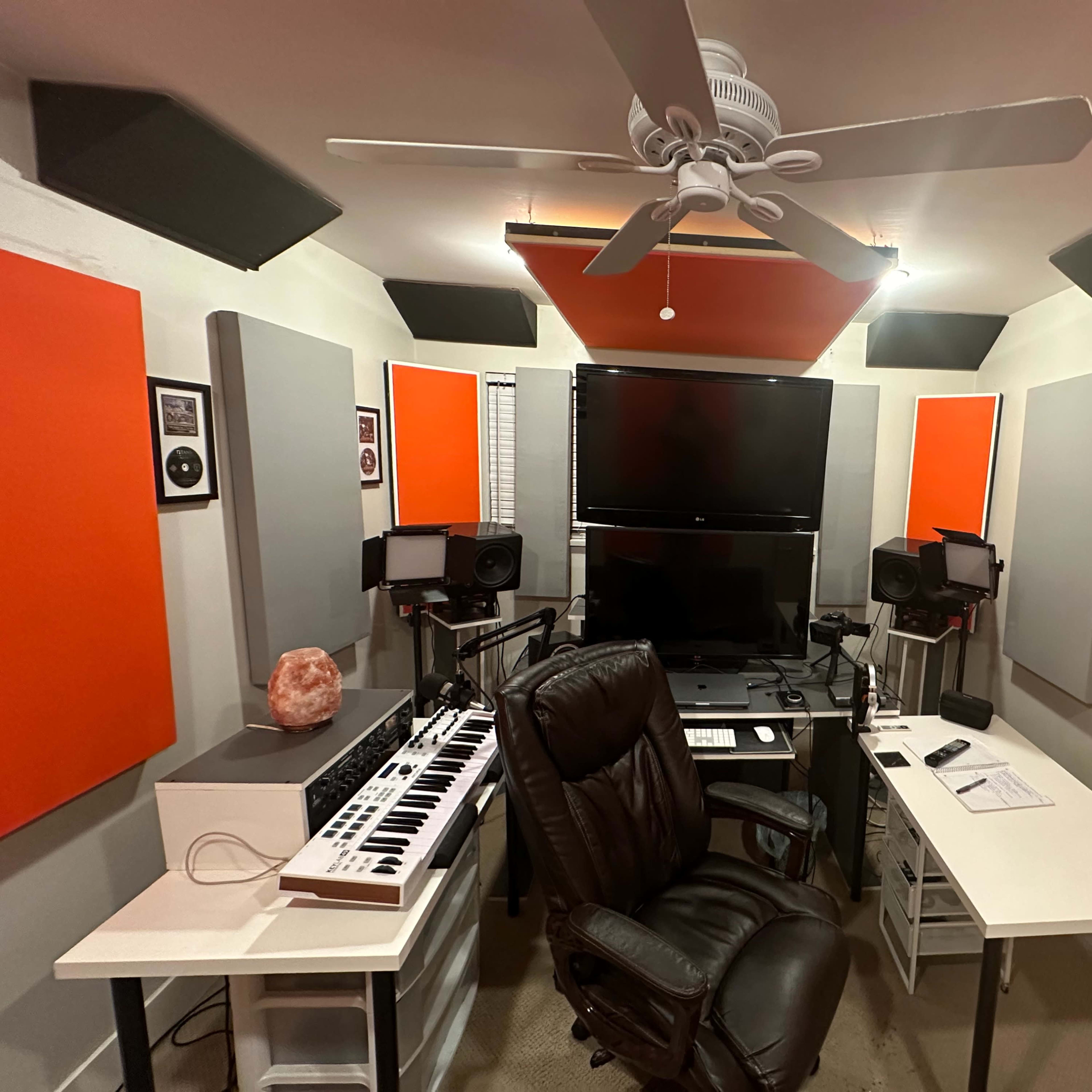 A recording studio with a black chair, computer monitors, a MIDI keyboard, and orange acoustic panels on the walls.