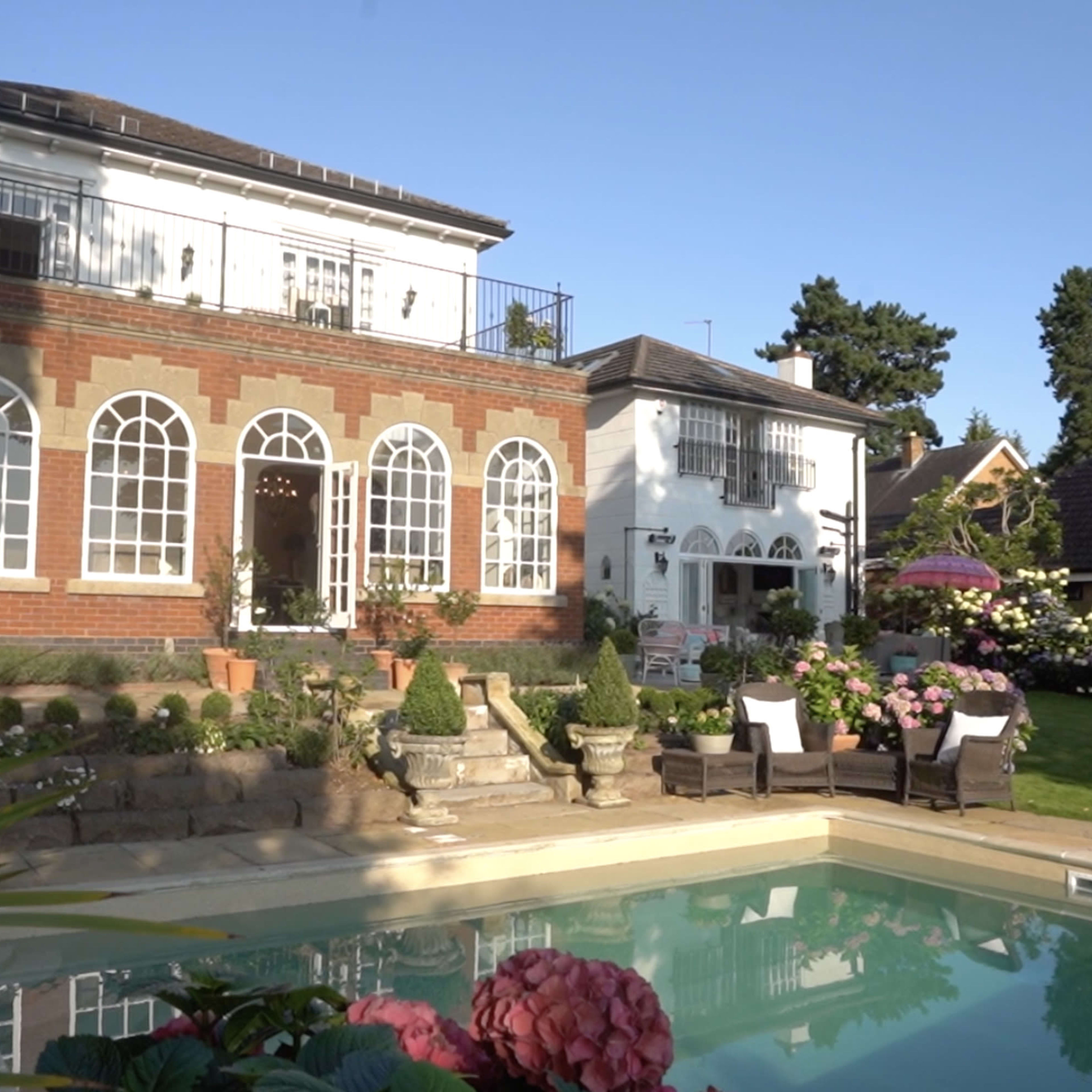 A modern house with large arched windows overlooks a swimming pool surrounded by meticulously landscaped gardens.
