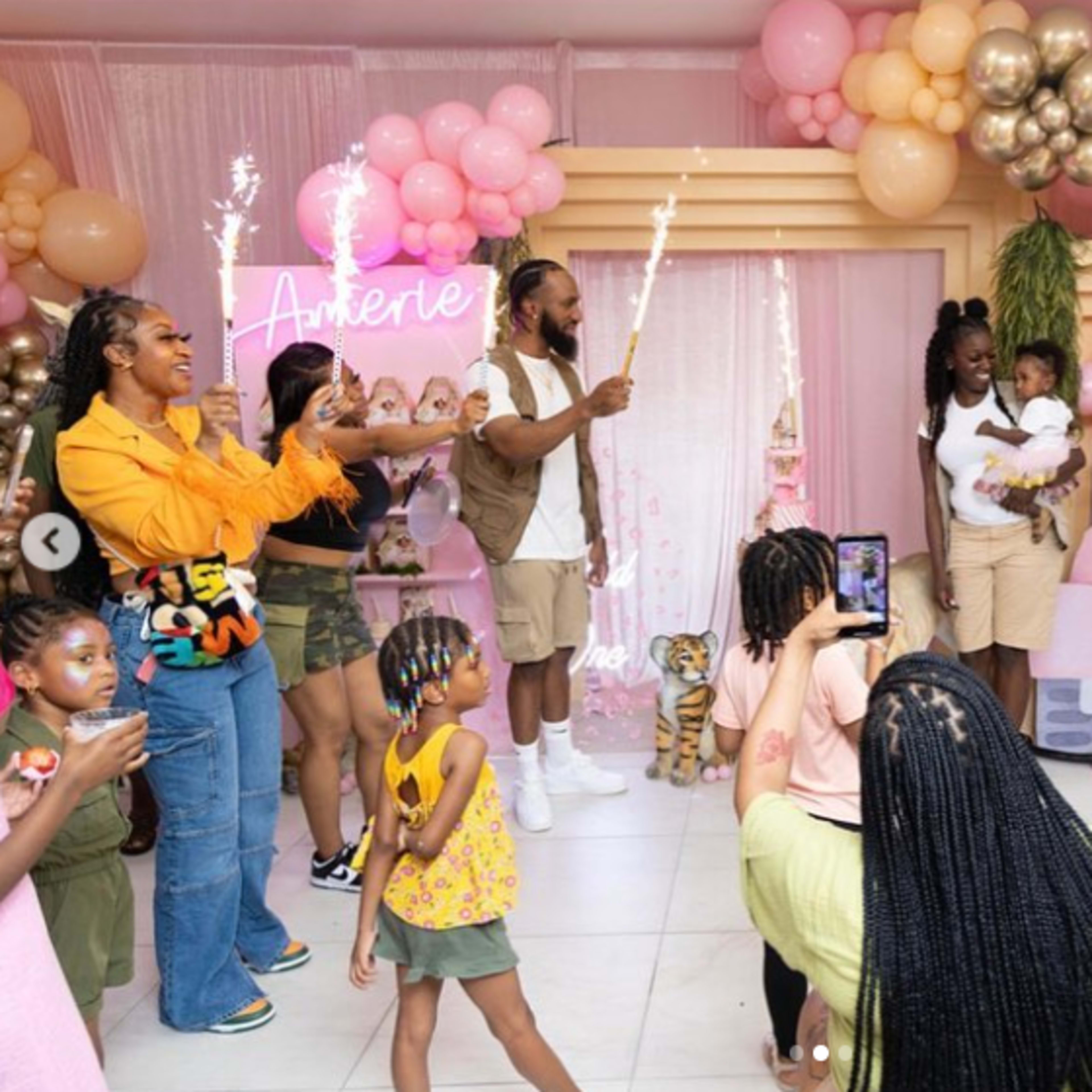 A group of people celebrates with sparklers in a brightly decorated room filled with balloons and a cake display.