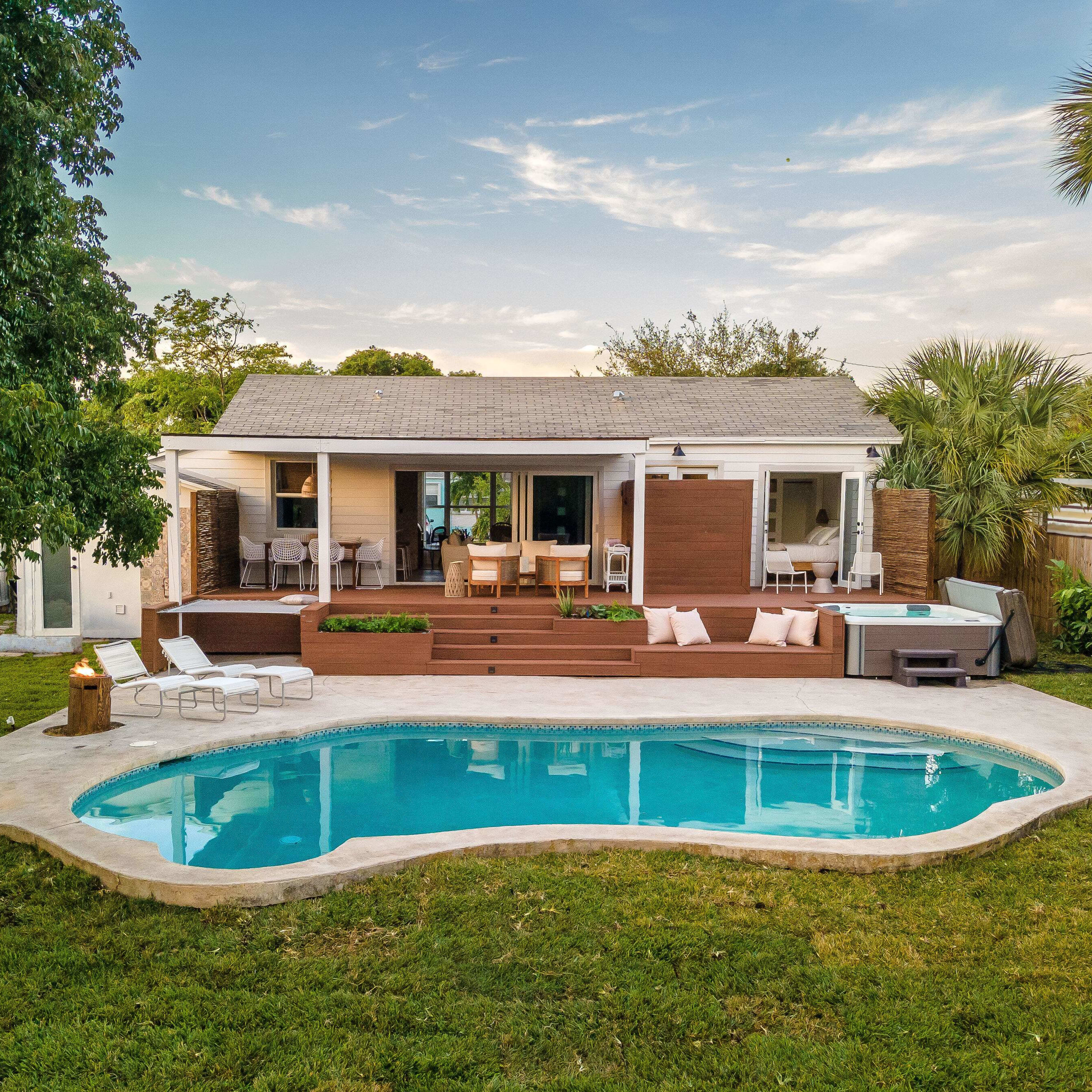 A single-story house with a backyard pool is surrounded by lawn furniture and palm trees.
