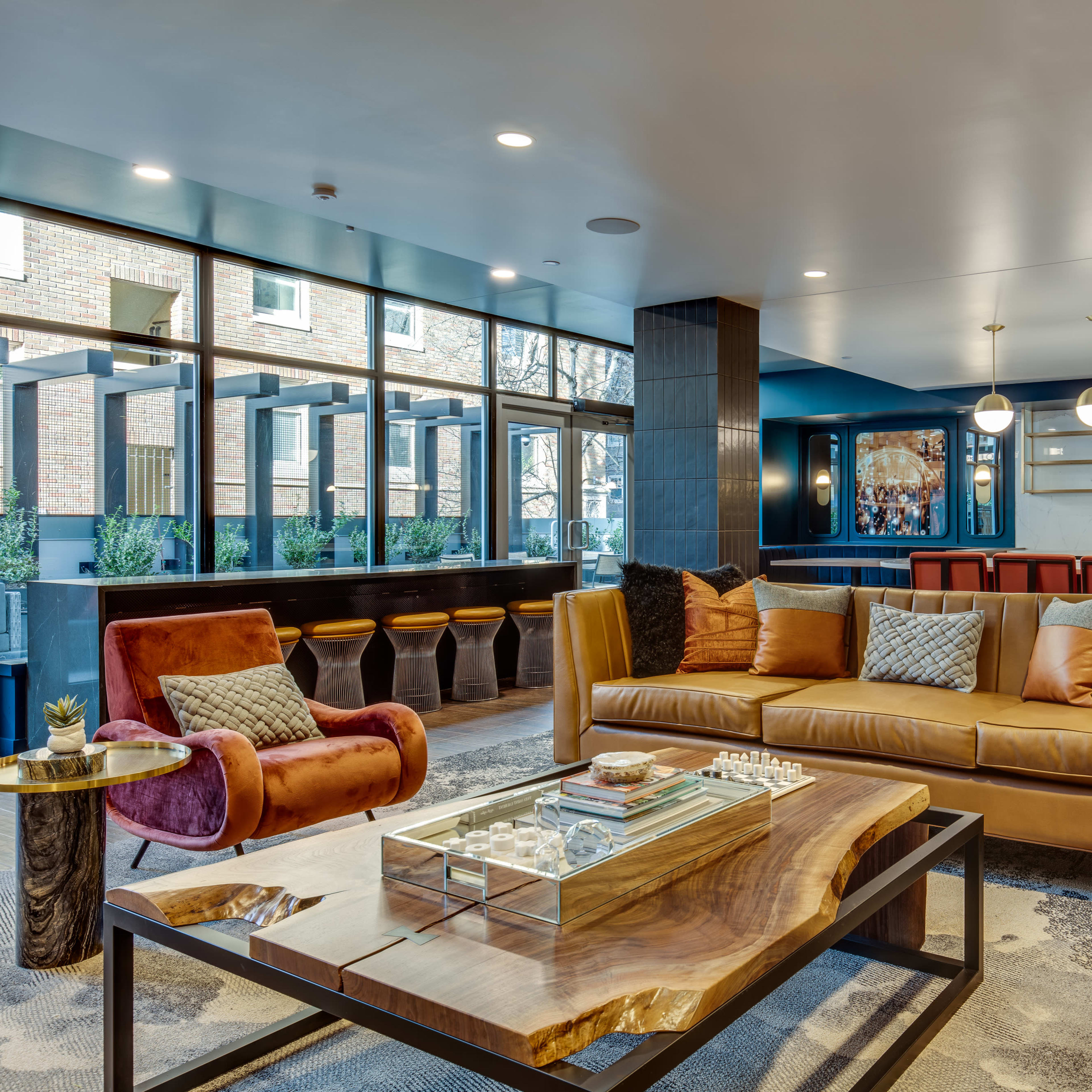 A modern lounge area featuring a brown leather sofa, a red accent chair, a wooden coffee table, and large windows letting in natural light.
