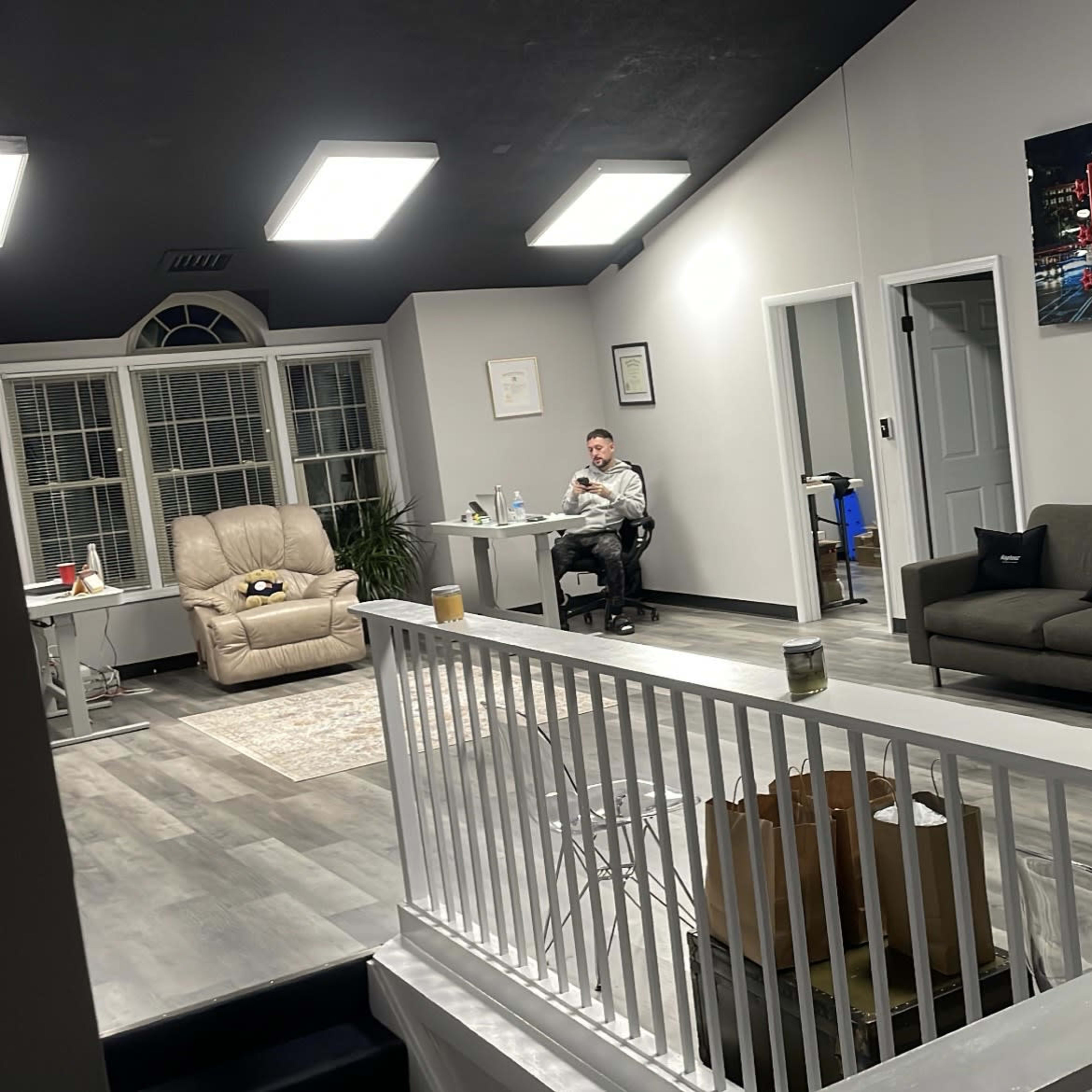A spacious, modern interior with a man working at a desk, a gray sofa in the center, and a large mirror on one wall.