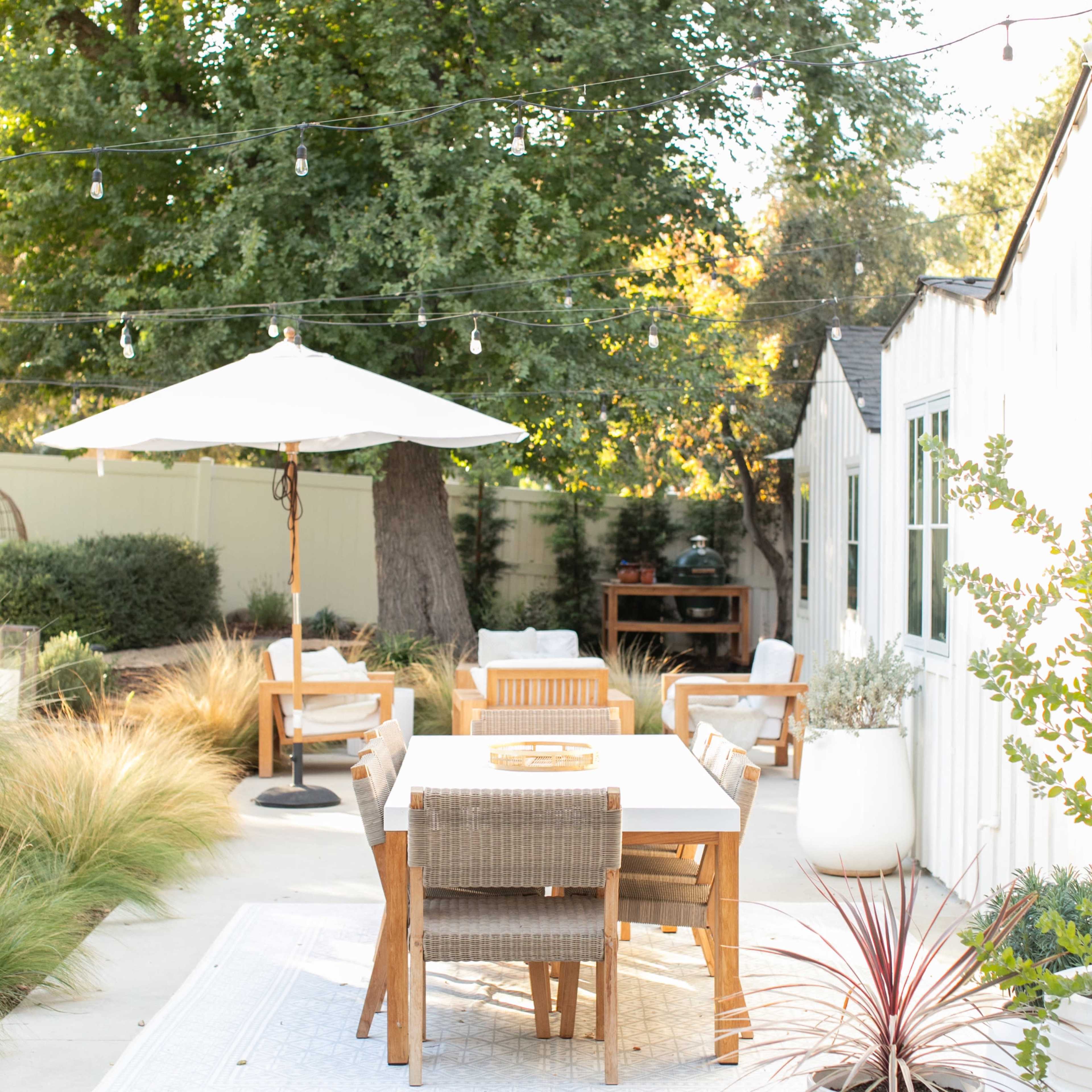 A patio dining area features a wooden table surrounded by chairs, an umbrella, and string lights, with greenery and a white house in the background.