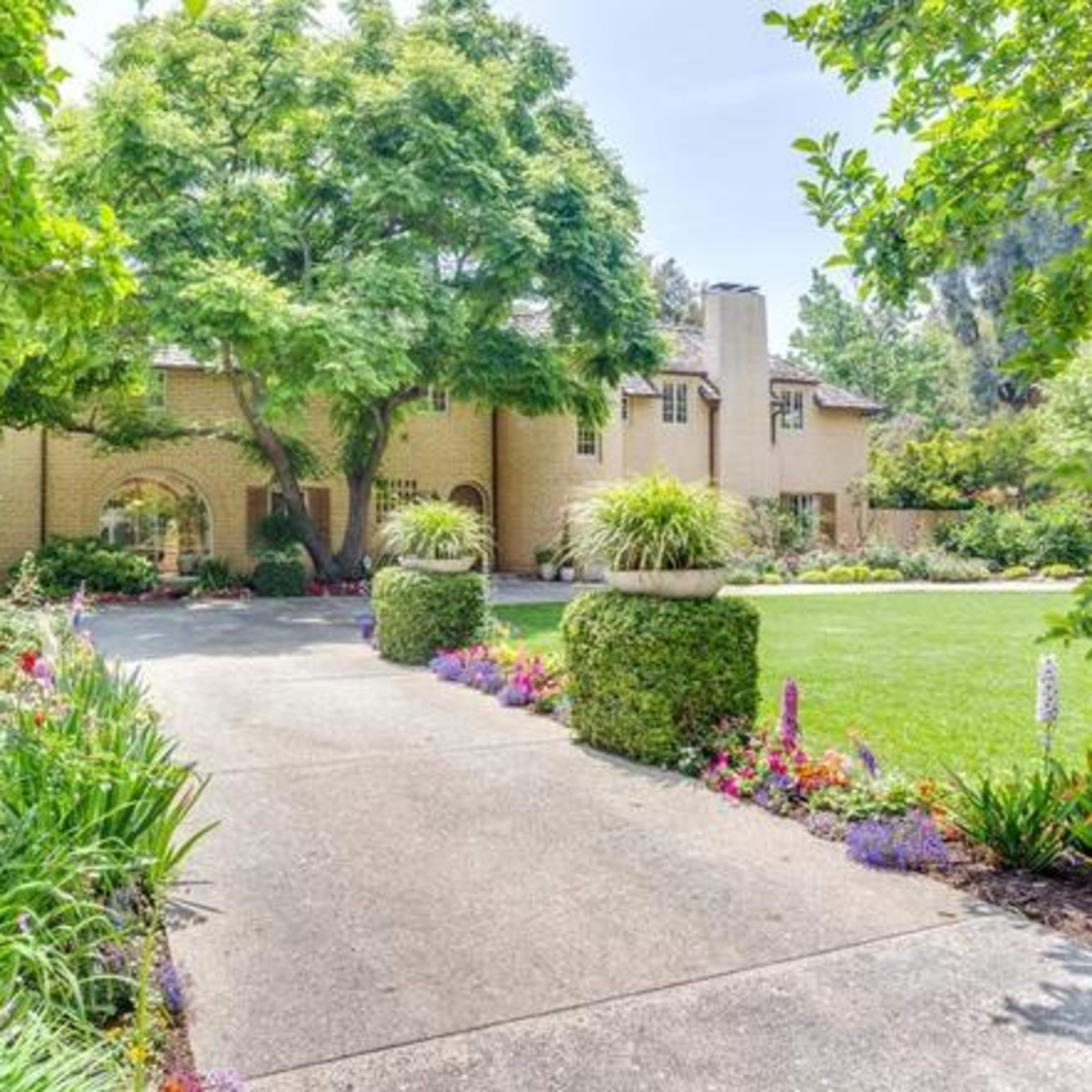 A landscaped driveway leads up to a two-story brick house surrounded by trees and colorful flower beds.