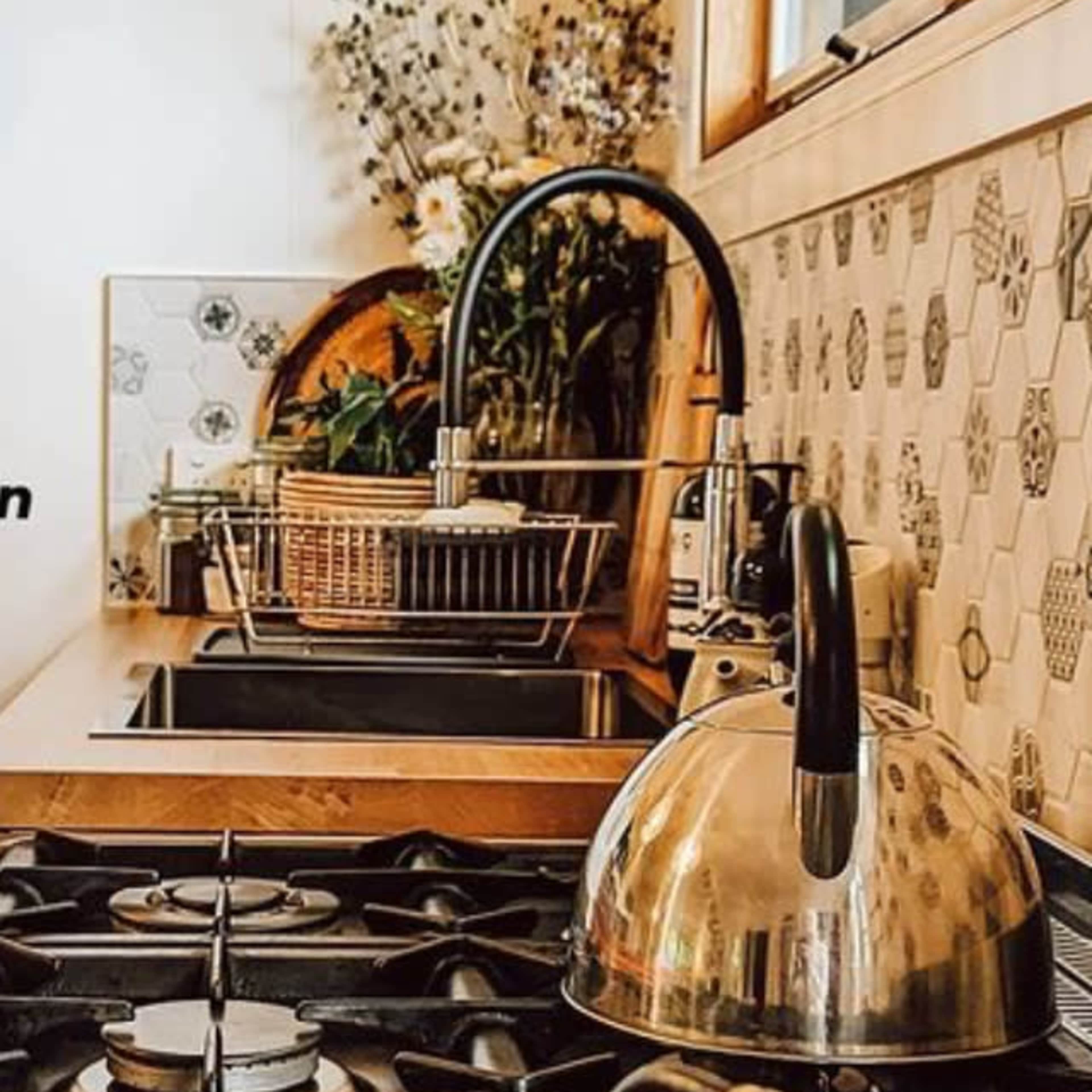 A kitchen countertop with a gas stove, a stainless steel kettle, a drying rack, and a vase of flowers in the background.