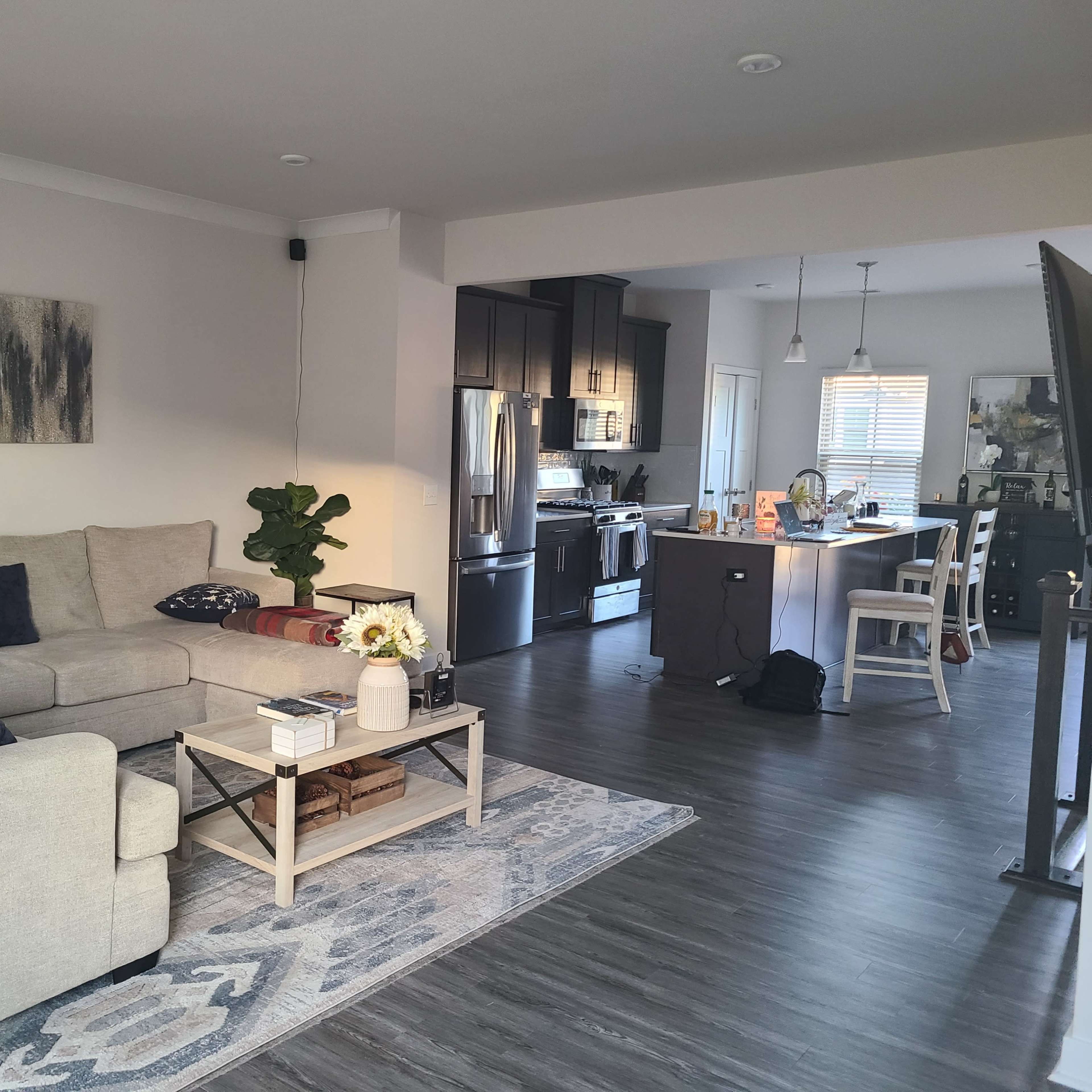 A living room with a light-colored sectional sofa, a coffee table, and an adjacent kitchen area featuring dark cabinetry and stainless steel appliances.