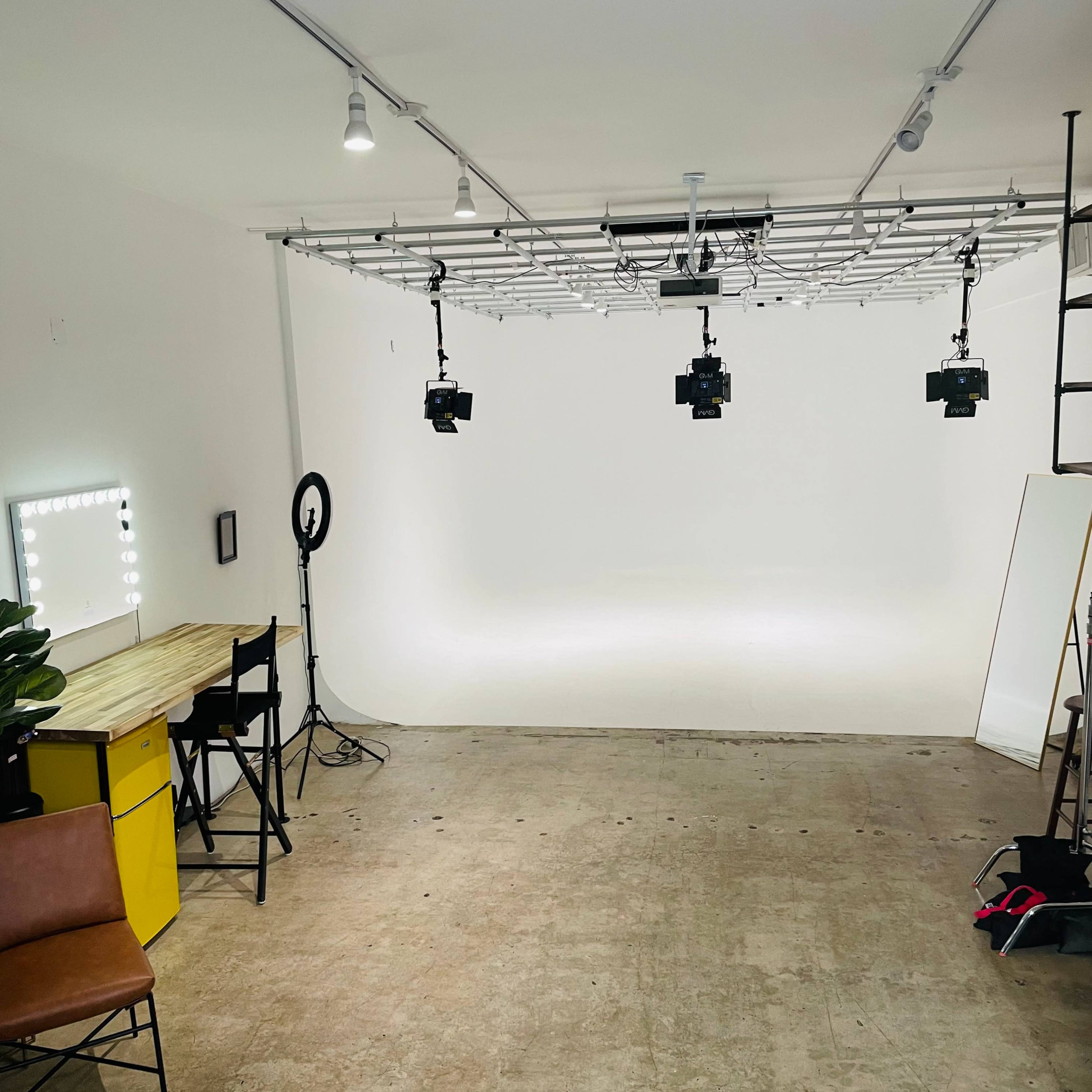 A minimalist studio space with a white backdrop, multiple lighting setups, a desk, and a chair.