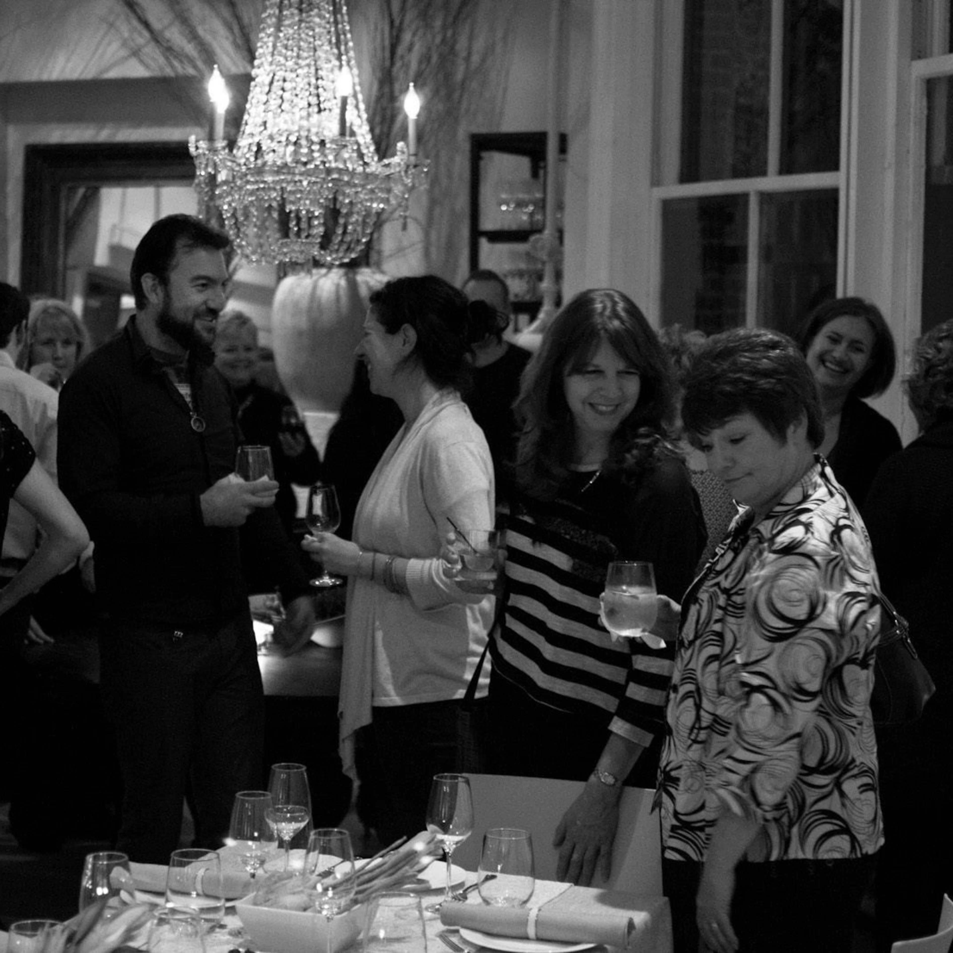 A group of people converse and socialize in a dimly lit dining room adorned with a chandelier.