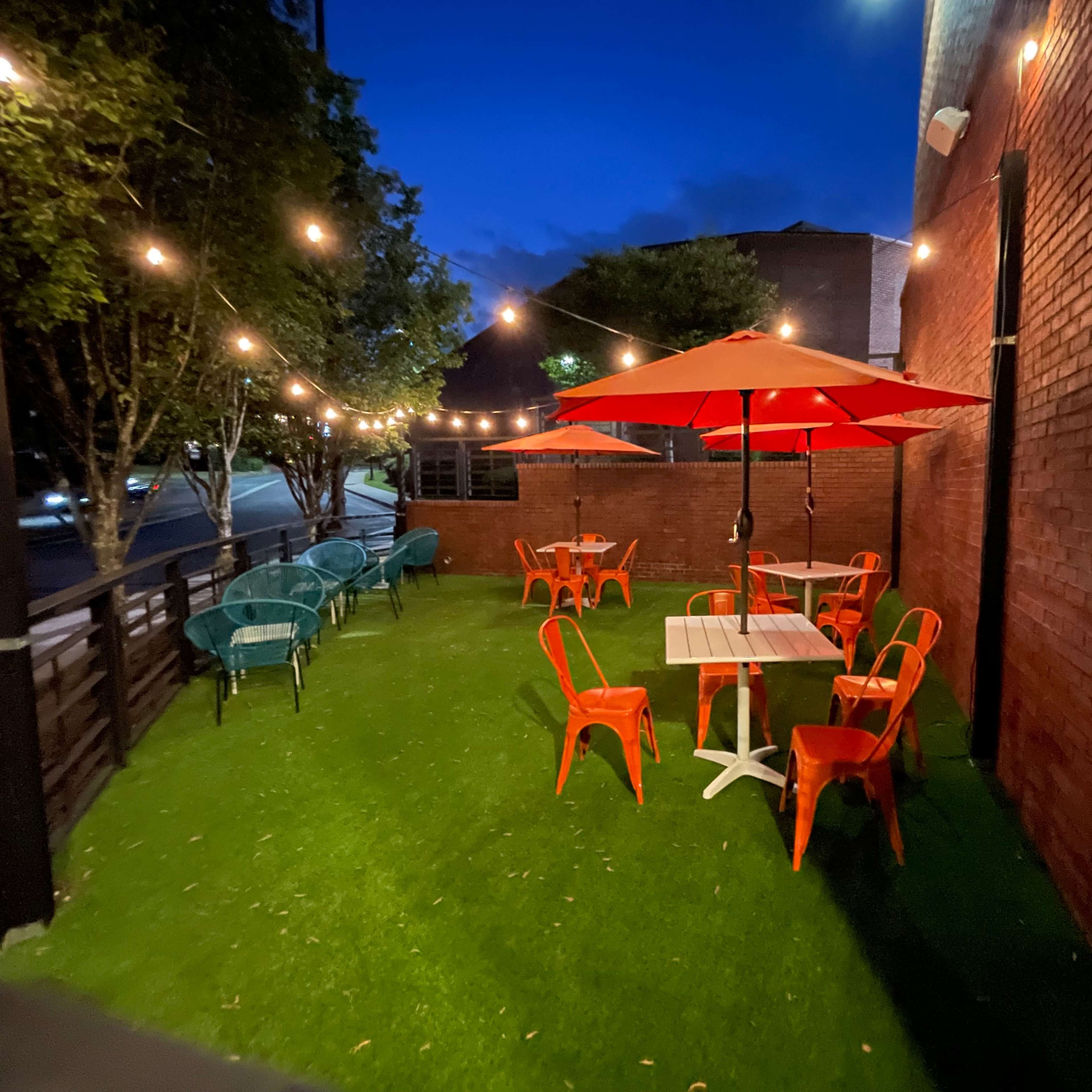 An outdoor seating area with orange chairs and umbrellas on a grassy surface, illuminated by string lights against a twilight sky.