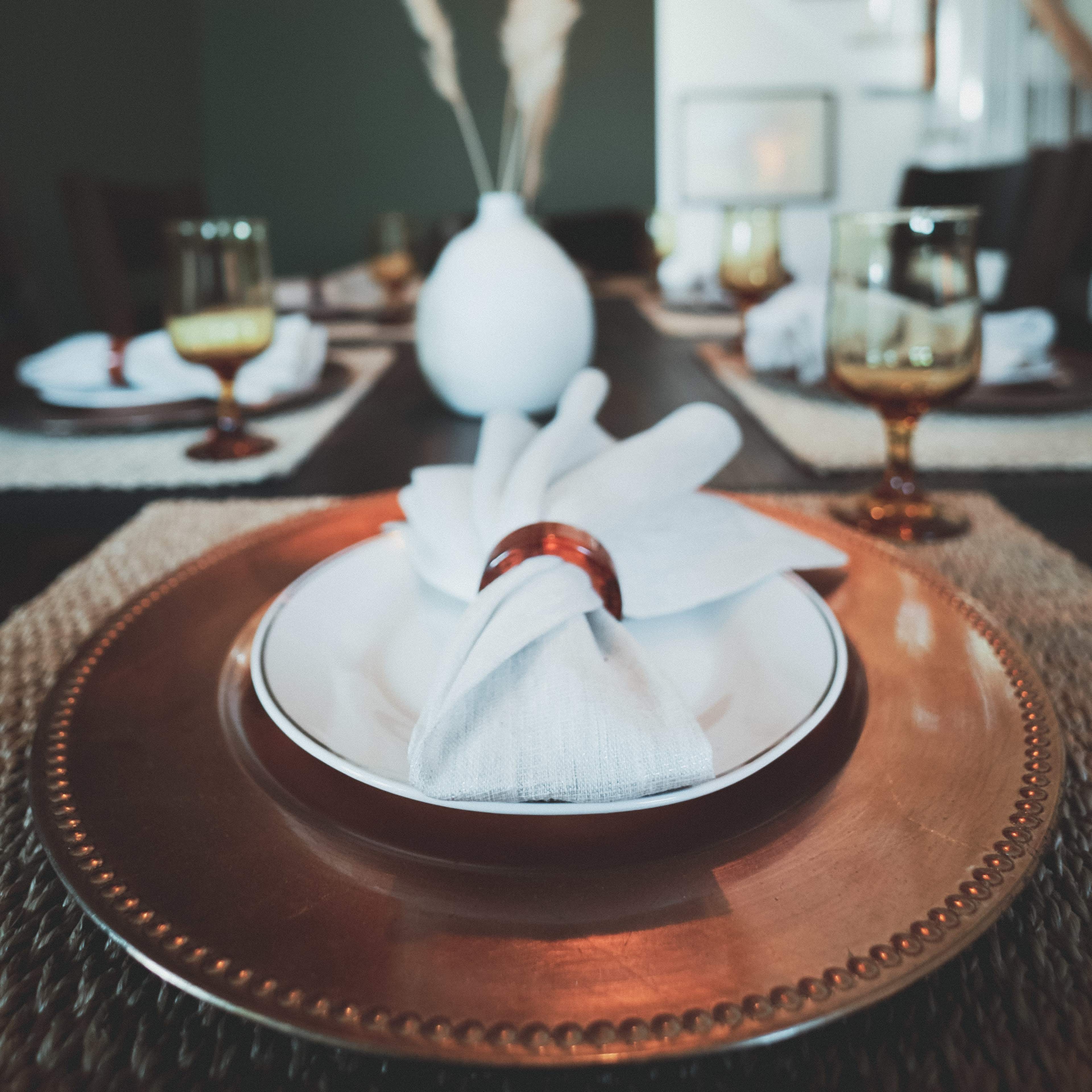 A dining table is set with white plates, copper chargers, and decorative napkins, accompanied by glassware and a simple vase with dried plants.