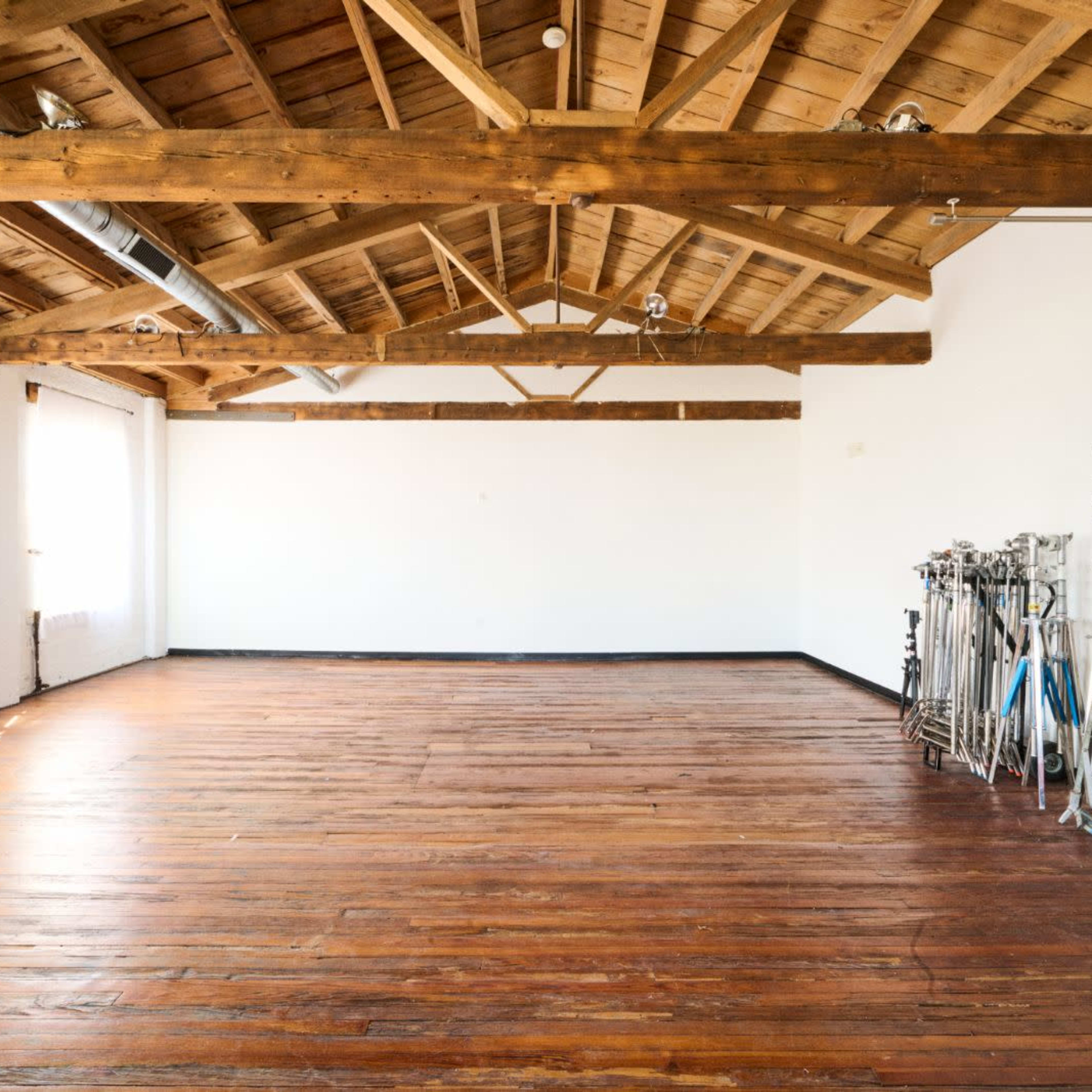 A spacious room with wooden beams and a polished wooden floor, featuring large windows and equipment stands along one wall.