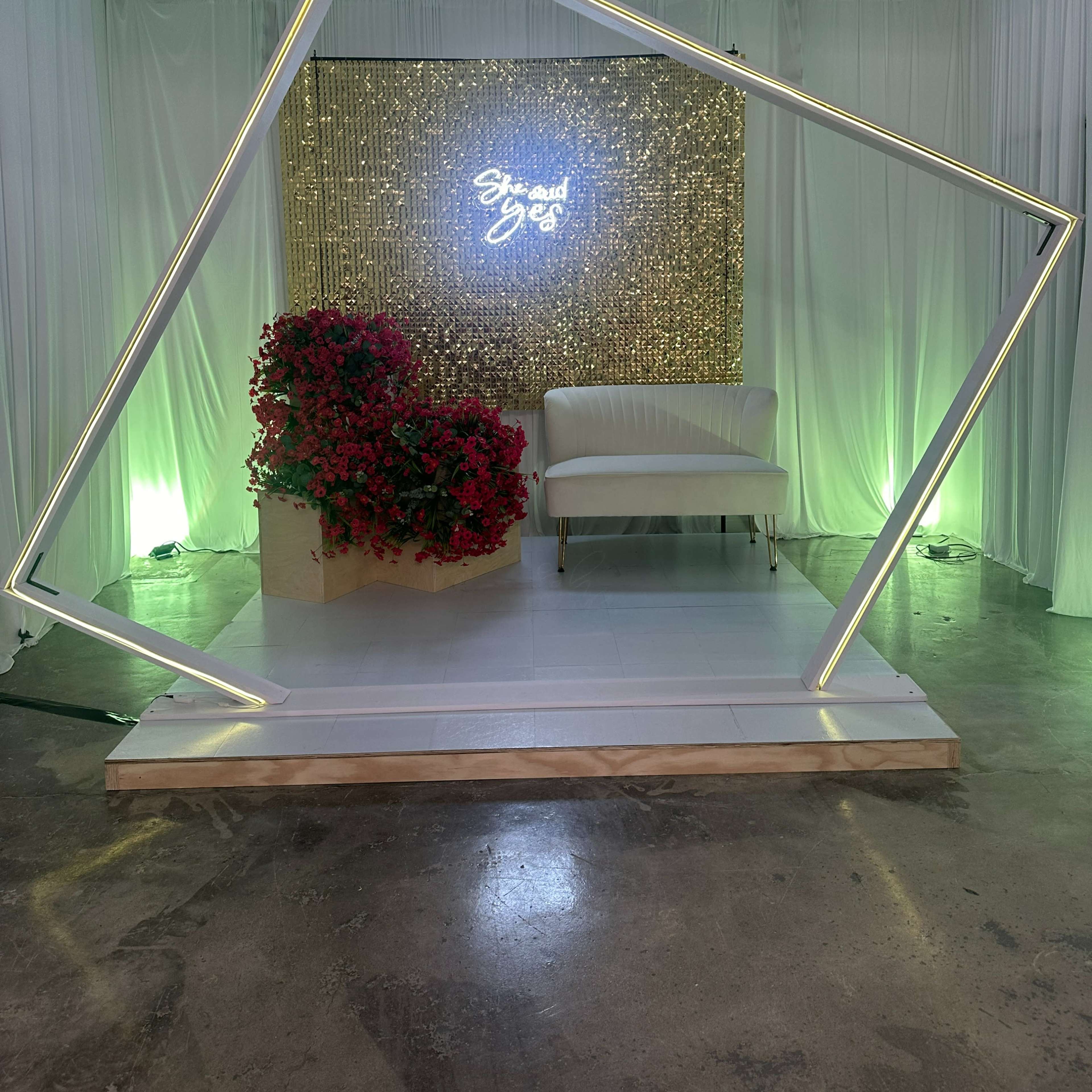The scene features a modern, illuminated photo backdrop with a neon sign, surrounded by white drapes, a floral arrangement, and a white couch.