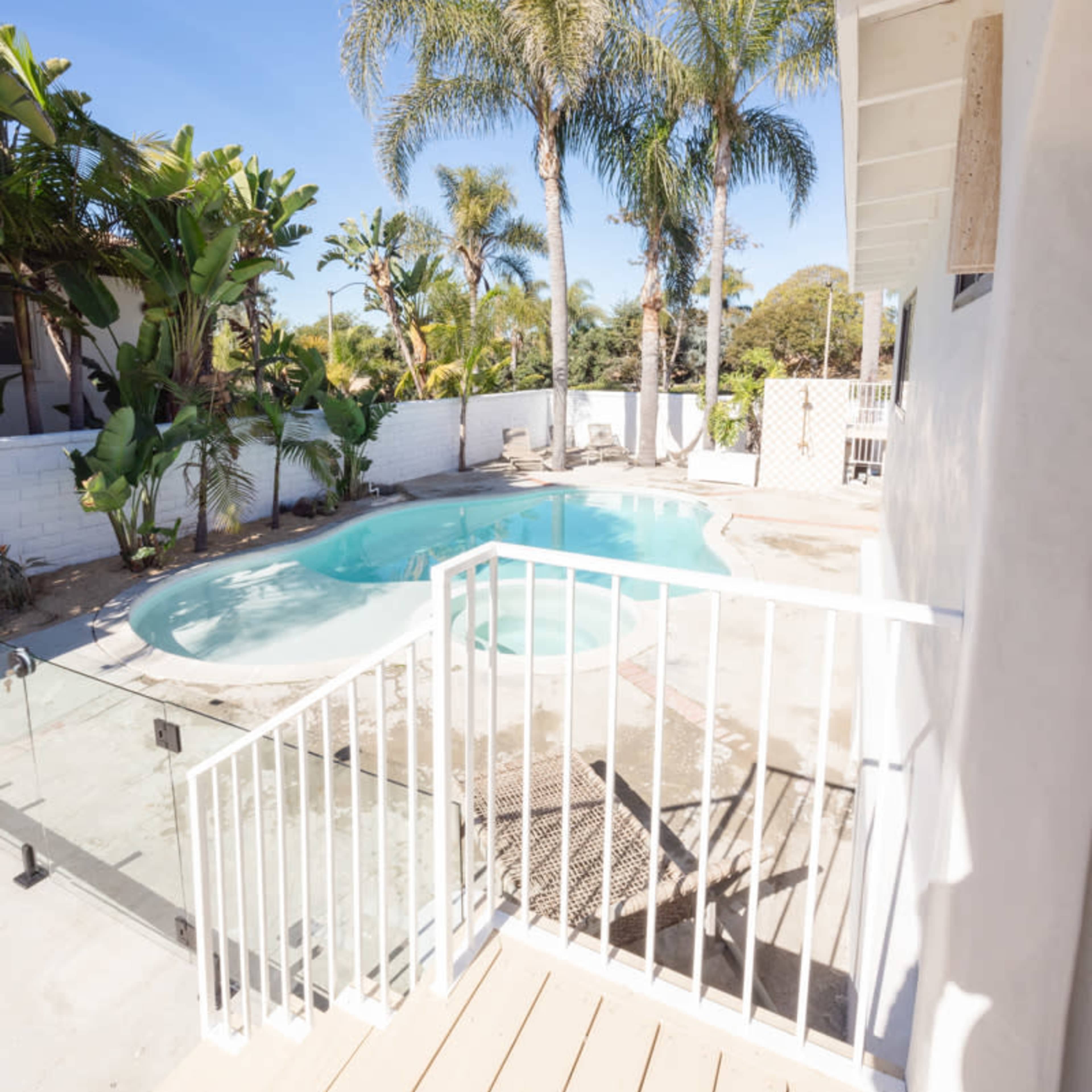 A backyard with a swimming pool surrounded by palm trees and plants, with a white railing and wooden deck in view.