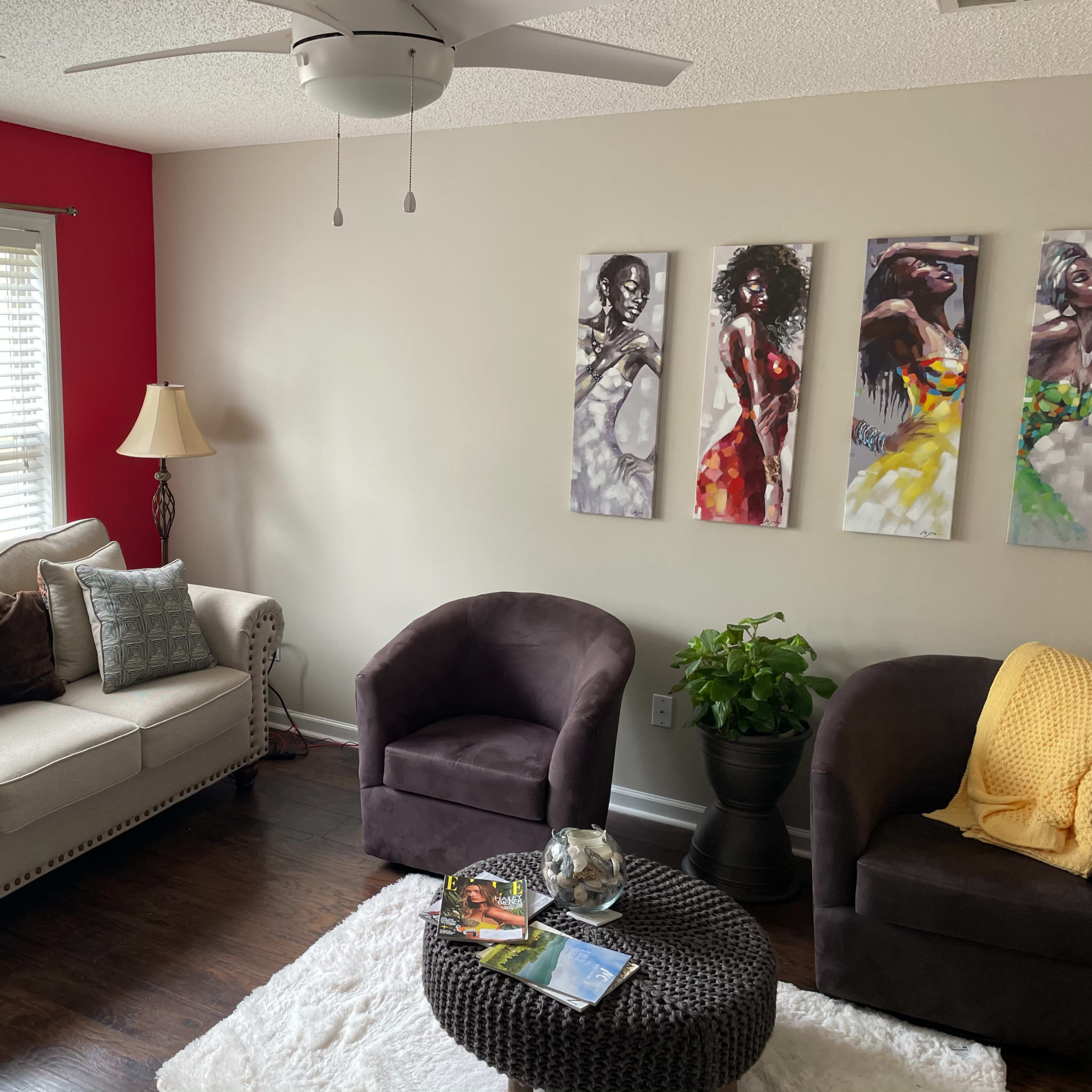 The living room features a light-colored sofa with decorative cushions, two dark armchairs, a round coffee table with books and a plant, and three paintings on the wall, with a red accent wall behind.