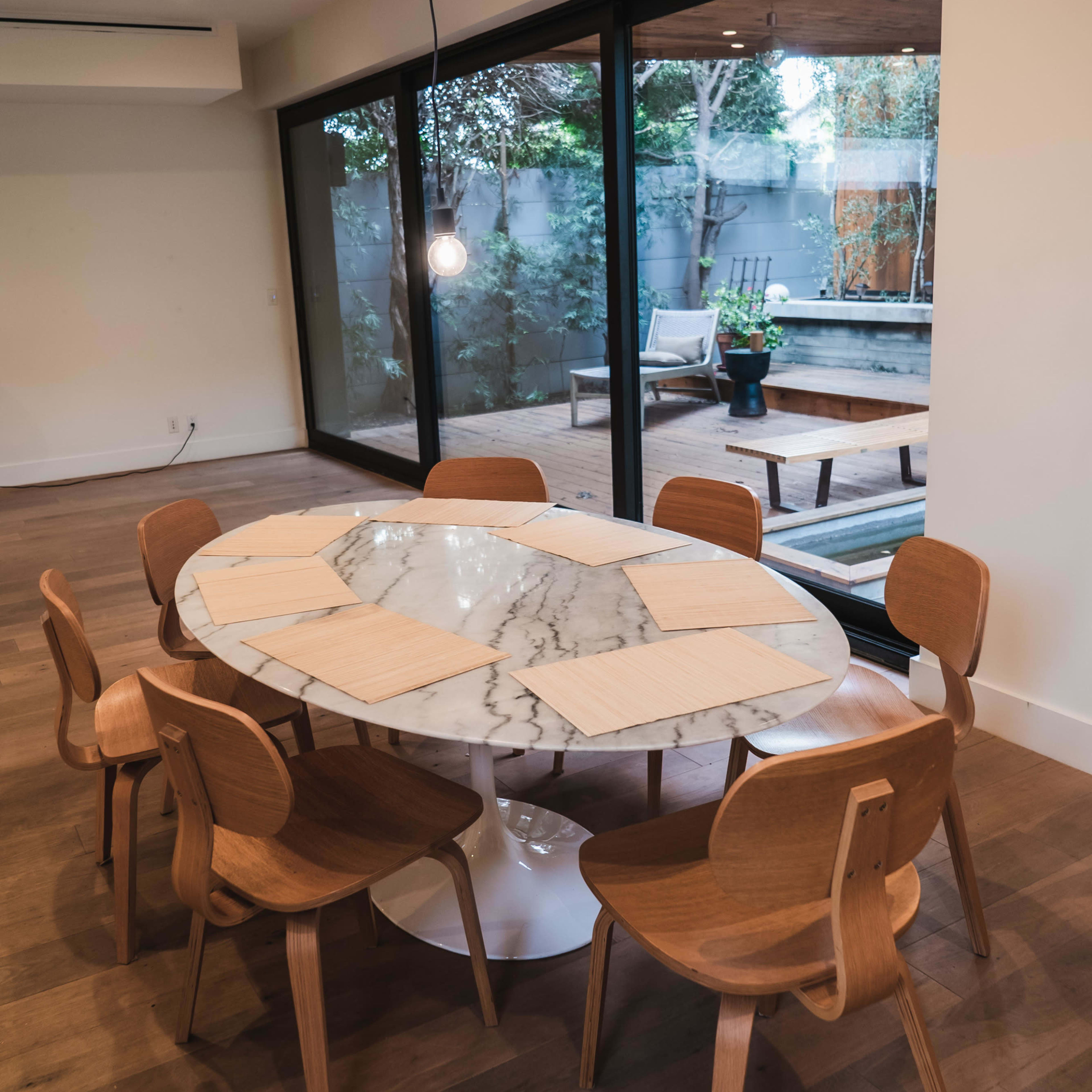 A round marble dining table with six wooden chairs is positioned in a room that features large glass doors opening to a patio view.