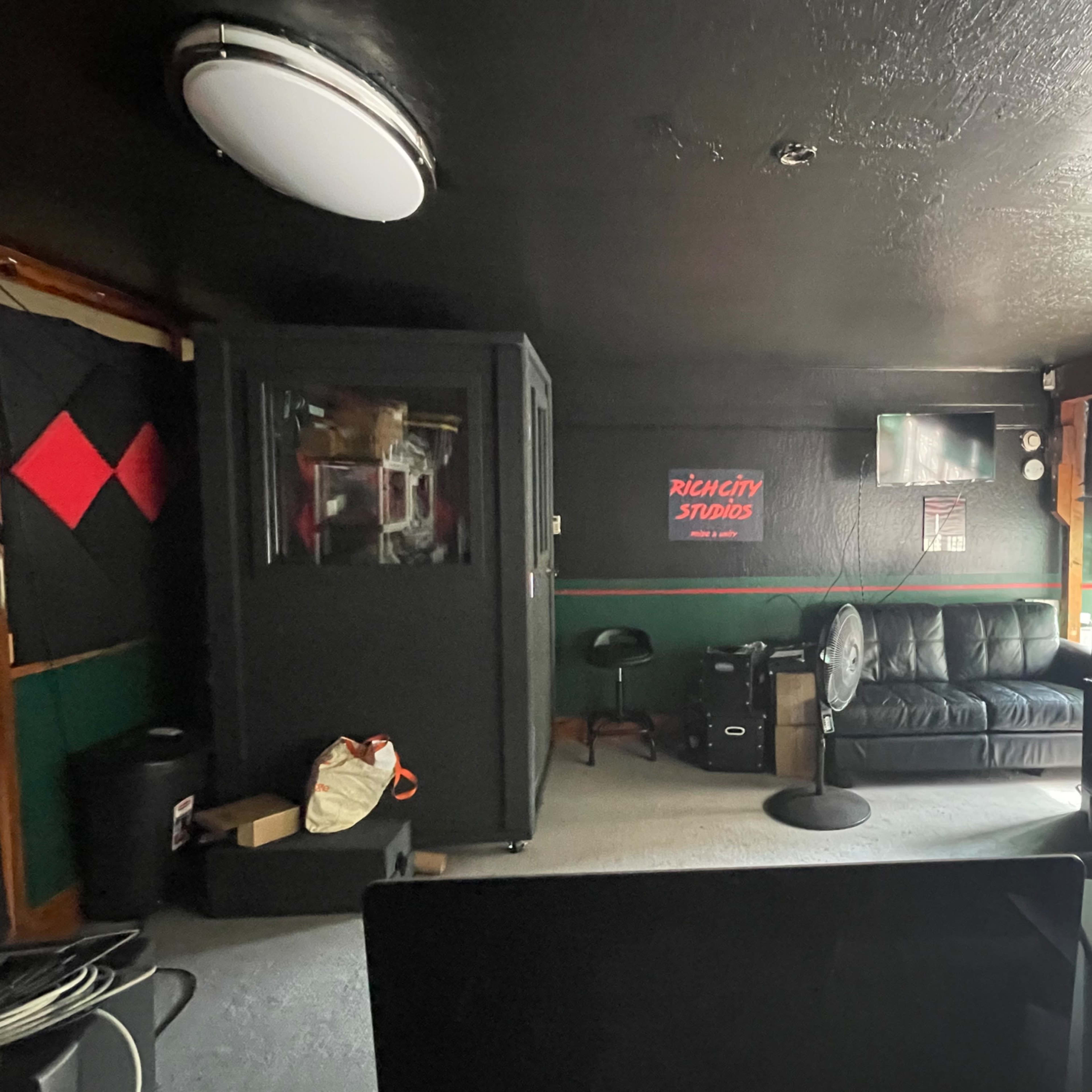A dimly lit room with a black leather couch, a fan, and a soundproof booth along with a TV on the wall.