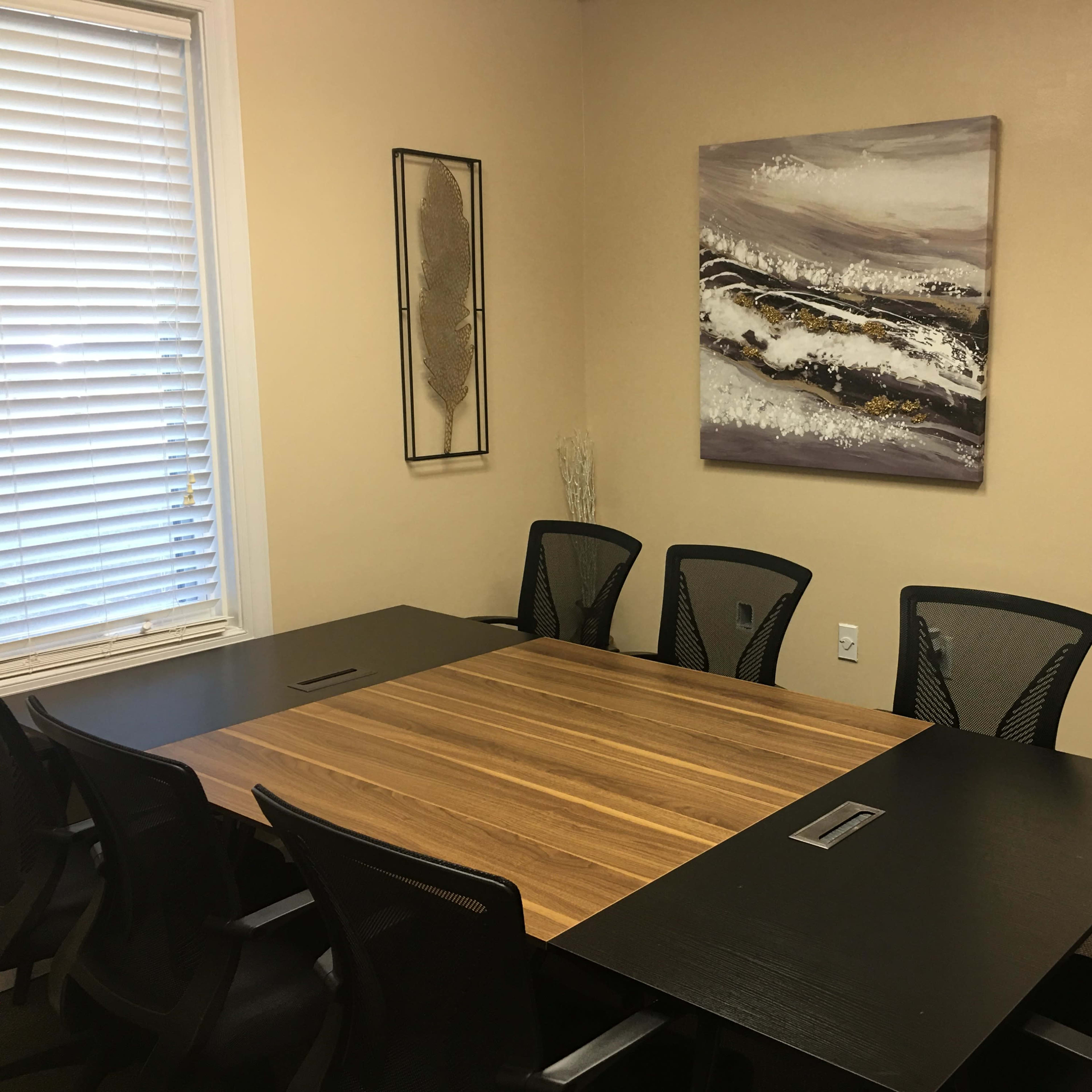 A conference room features a large wooden table surrounded by black mesh chairs, with windows allowing natural light and a landscape painting on the wall.
