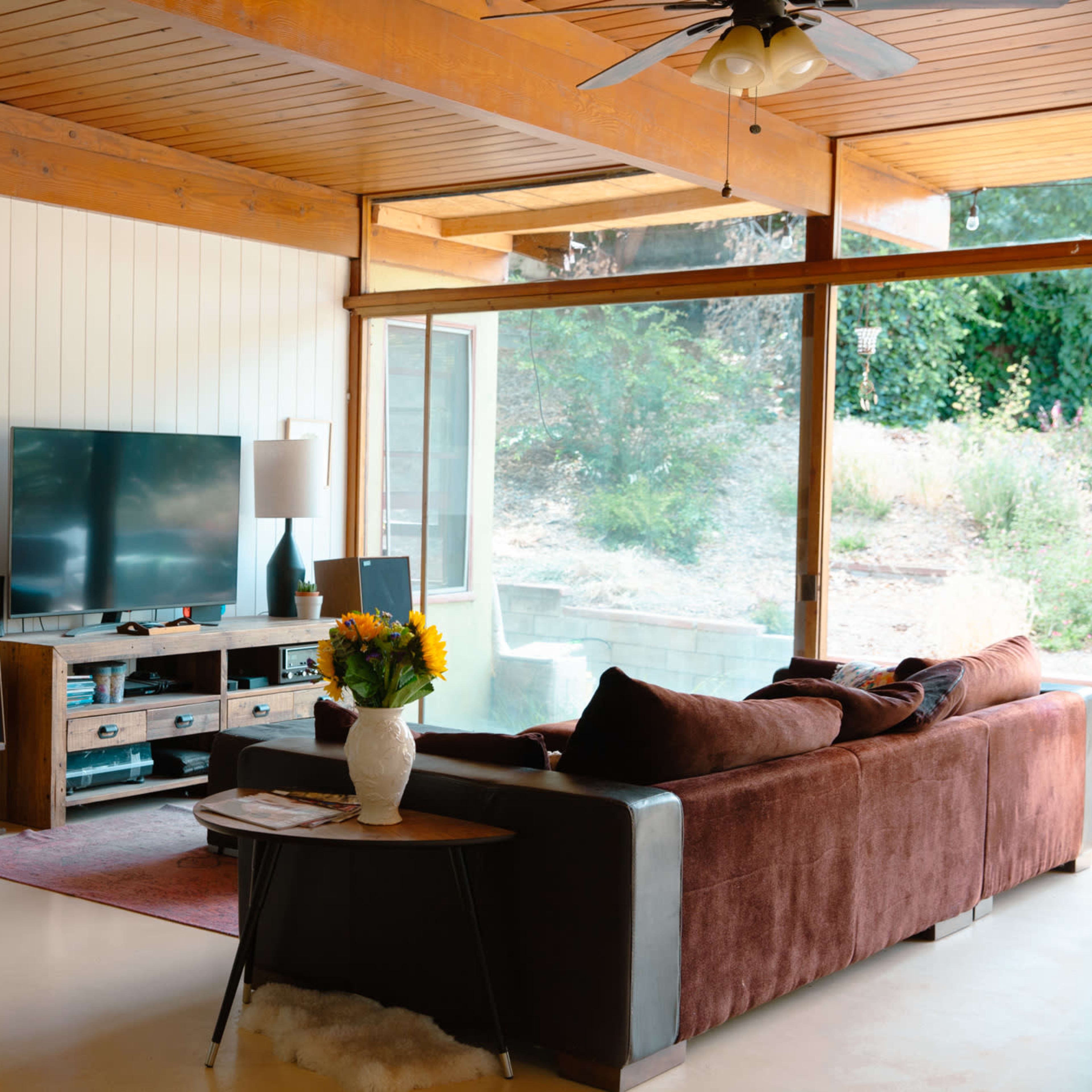 A living room features a brown sofa facing a TV unit, with large windows allowing natural light and a view of the outdoors.