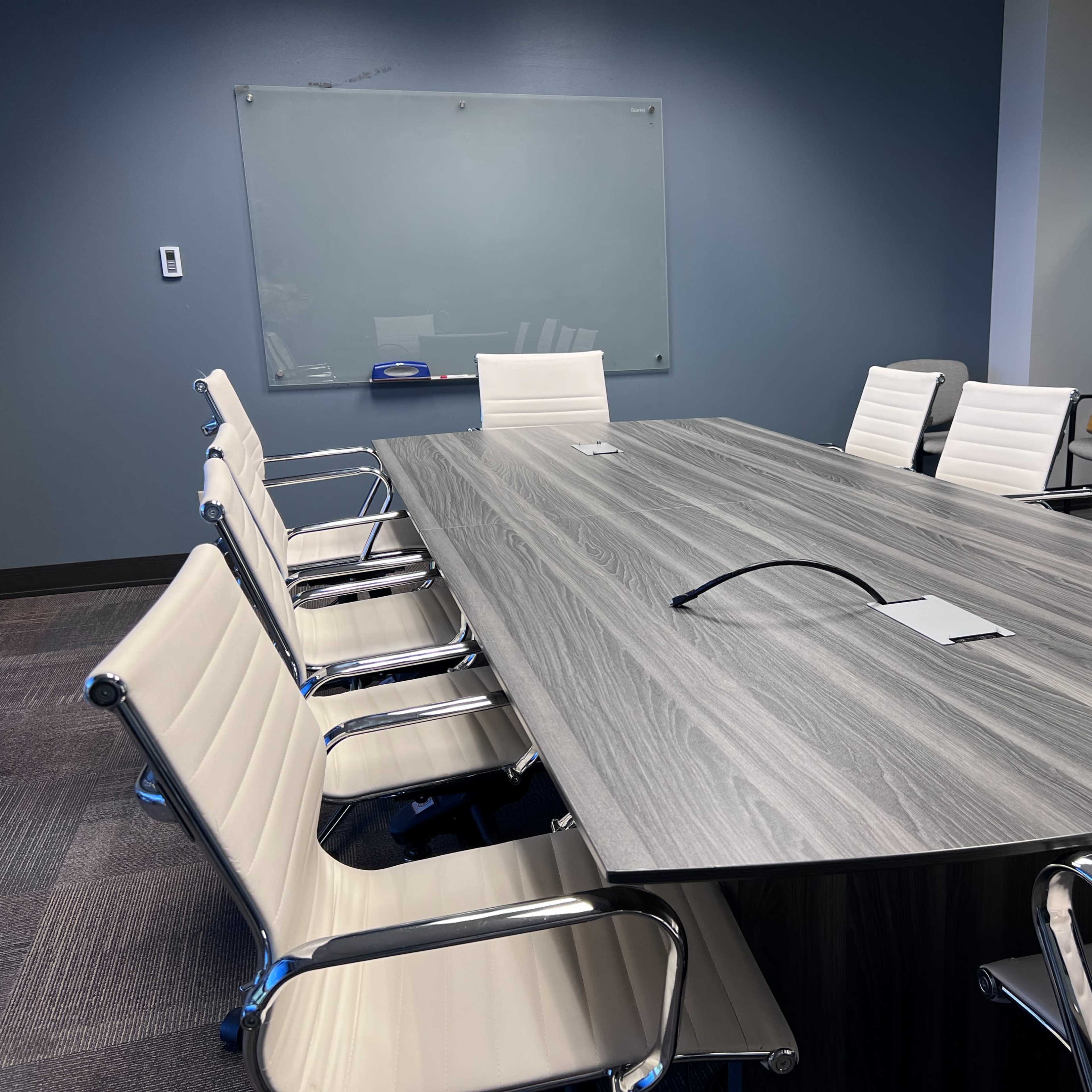 A modern conference room features a long gray table surrounded by white chairs, with a whiteboard visible in the background.