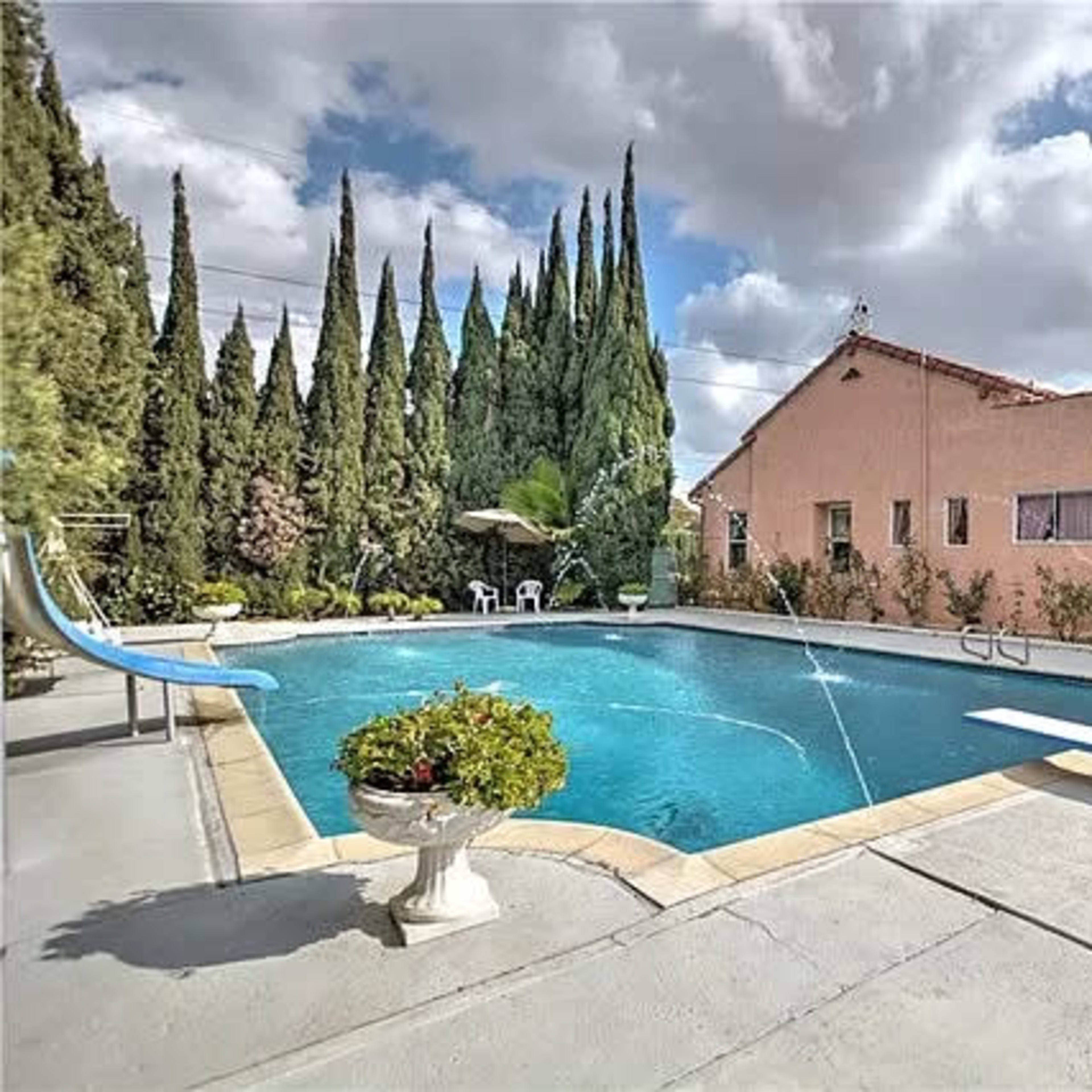 A residential backyard featuring a swimming pool with a slide, surrounded by tall trees and a single-story house.