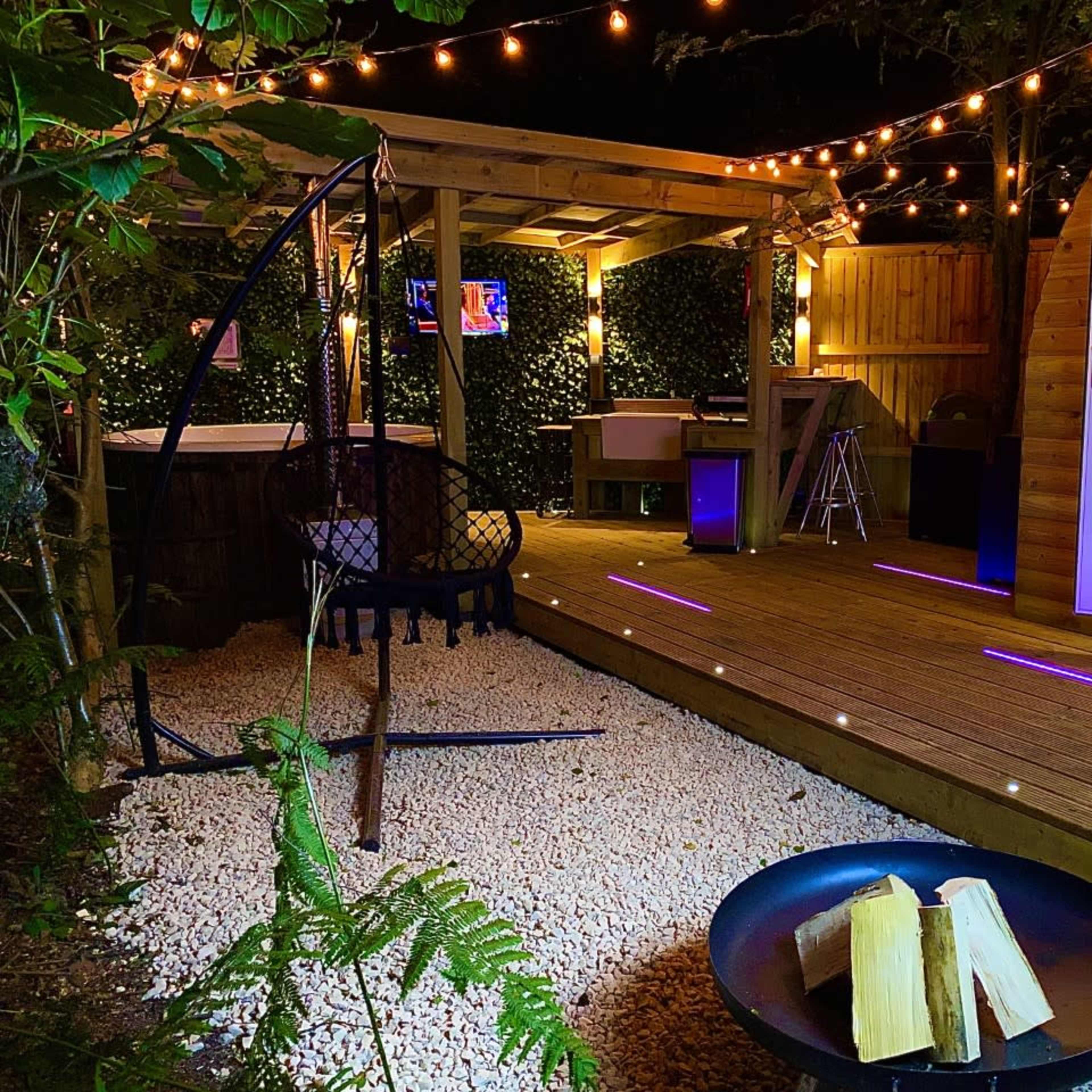 A well-lit outdoor deck area featuring a hanging chair, a fire pit with logs, and a cozy cabin-style room with double doors, surrounded by greenery and string lights.