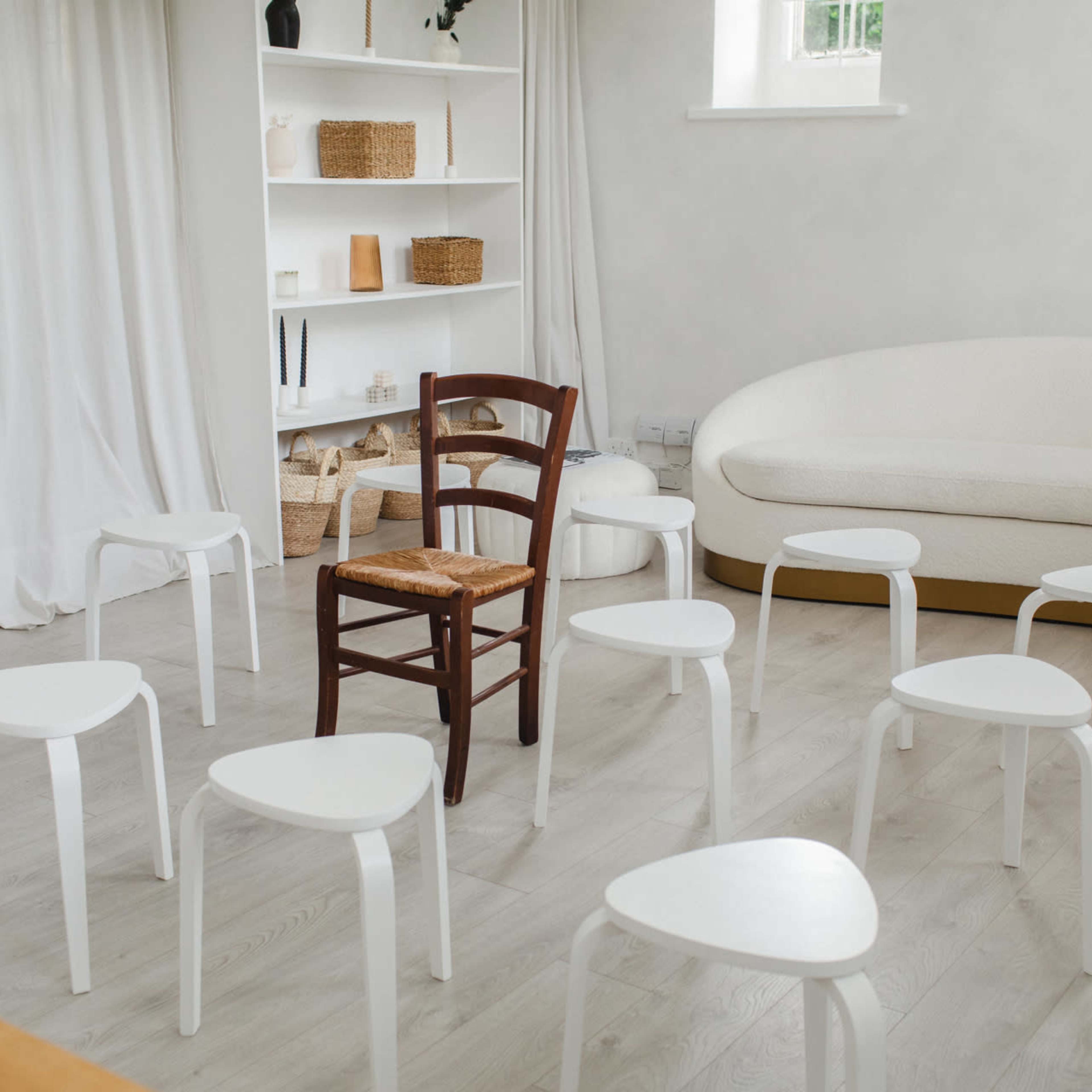A single wooden chair is placed among several white, triangular stools in a light-colored room with shelves and curtains.