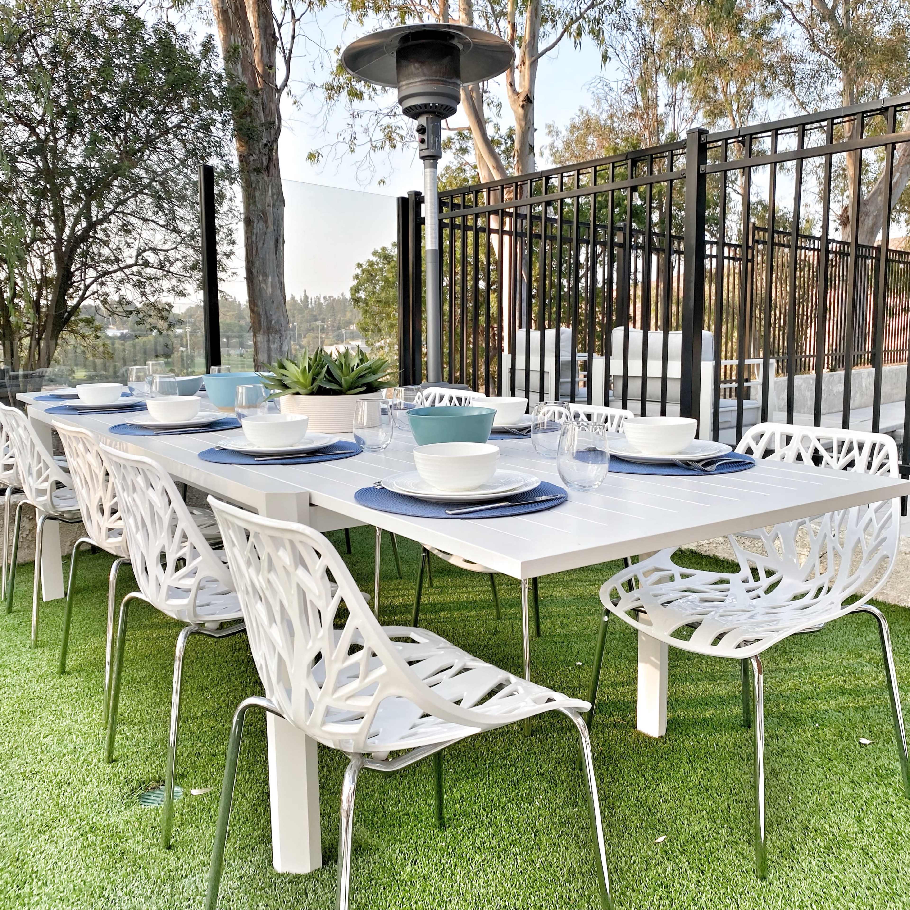 A long outdoor dining table is set for a meal, surrounded by decorative white chairs on green artificial grass, with a patio heater positioned overhead.
